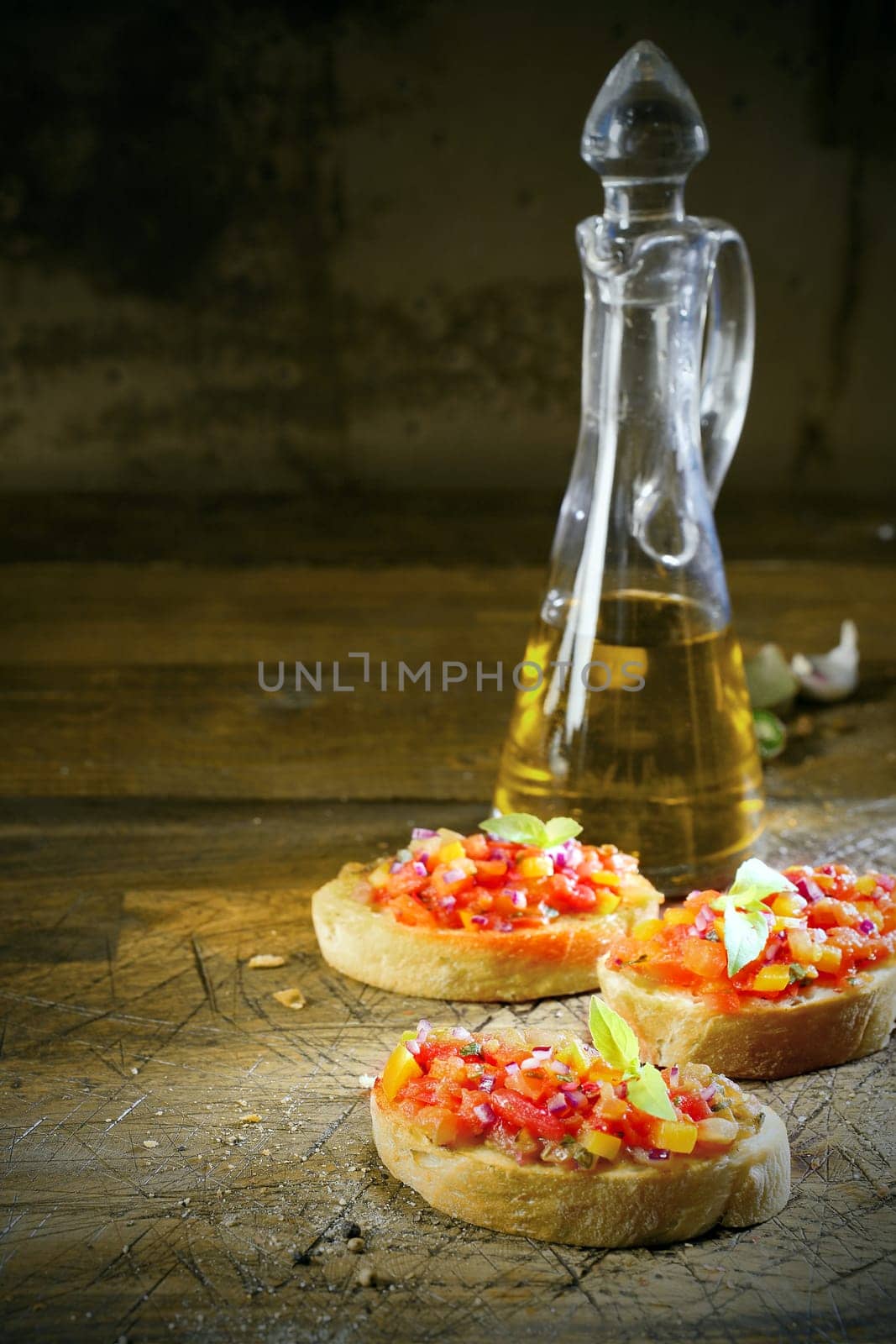 Preparing bruschetta in a country kitchen with three slices of toasted sliced baguette topped with chopped vegetables and oil with a decanter of virgin olive oil behind