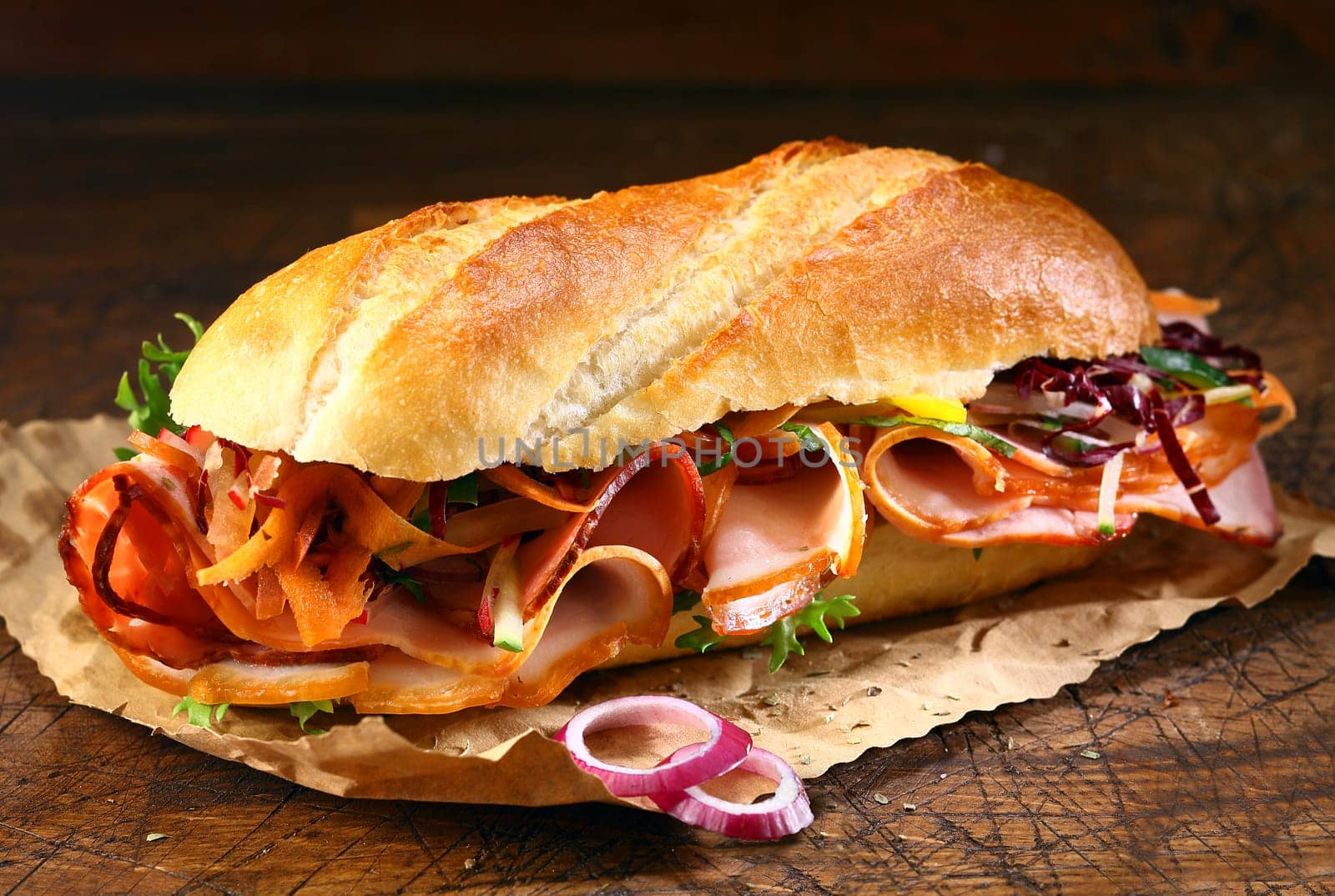 Delicious crusty baguette sandwich with ham, lettuce and onion on a small grungy crumpled sheet of brown paper on a wooden table
