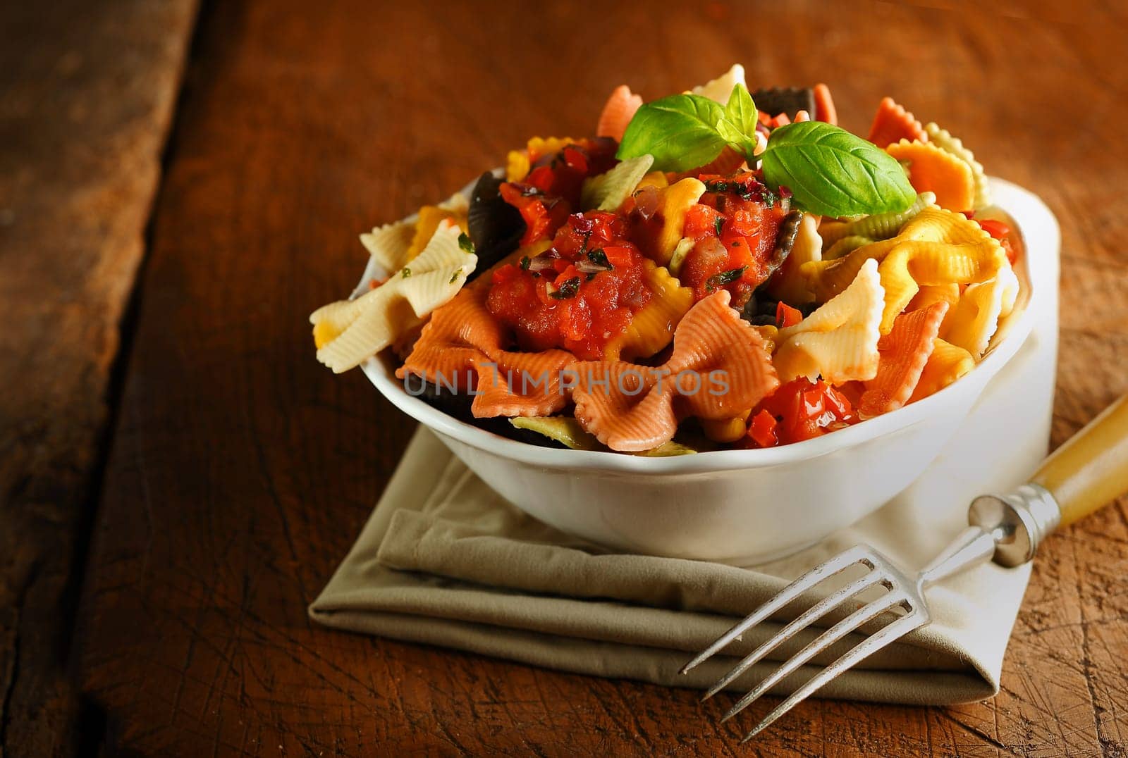 Bowl of cooked colourful Italian bow tie pasta topped with tomato sauce and basil standing on a folded napkin on an old wooden table with a fork