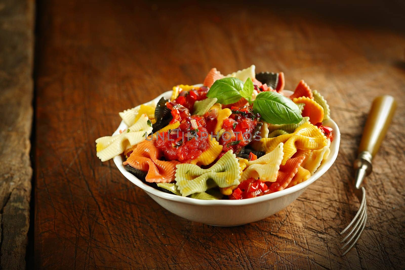 Multicoloured Italian bow tie pasta topped with tomato and garnished with basil served in a white ceramic bowl with a fork on an old wooden table