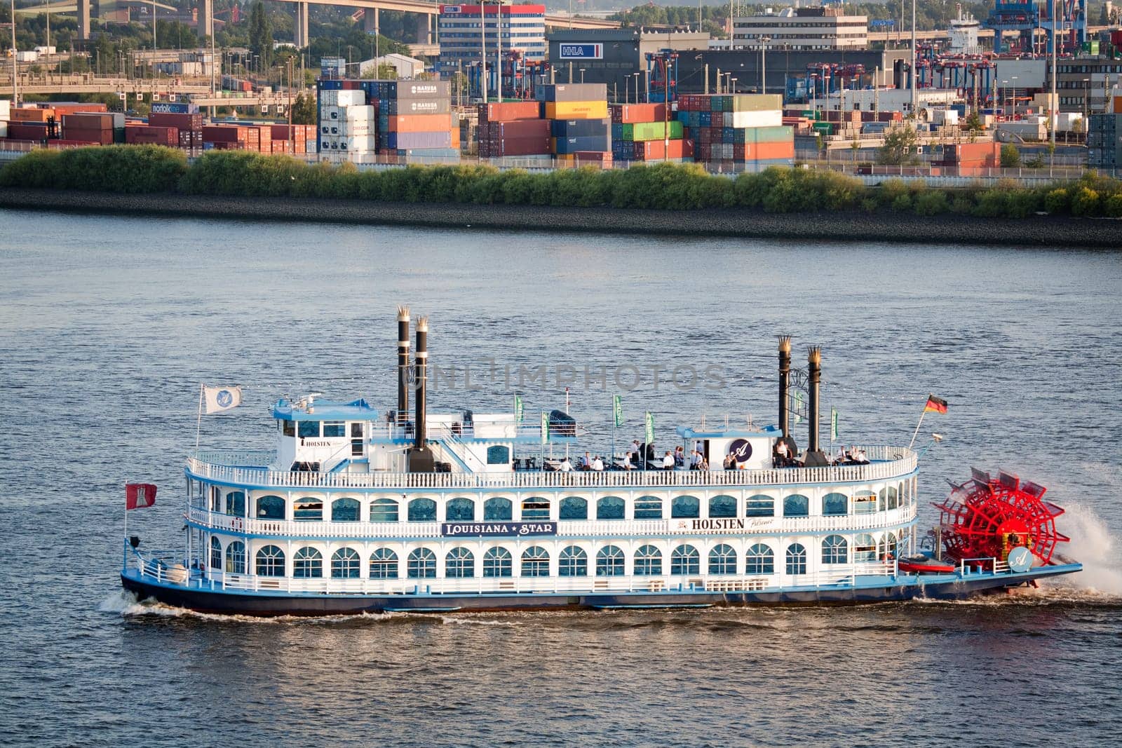 The paddle steamer Louisiana Star on the Elbe River in the Hamburg Harbor, Germany. Operated by the Louisiana Starboat Company, the boat is used for events and sightseeing primarily around the harbor area. The steamer was built in Germany to resemble the classic steamers one would associate with the Southern US, whereas the rear wheel motion is for aesthetic purposes only and the boat is driver by more conventional means. People can be seen on deck enjoying the view and Summer weather, with container terminals in the background.
