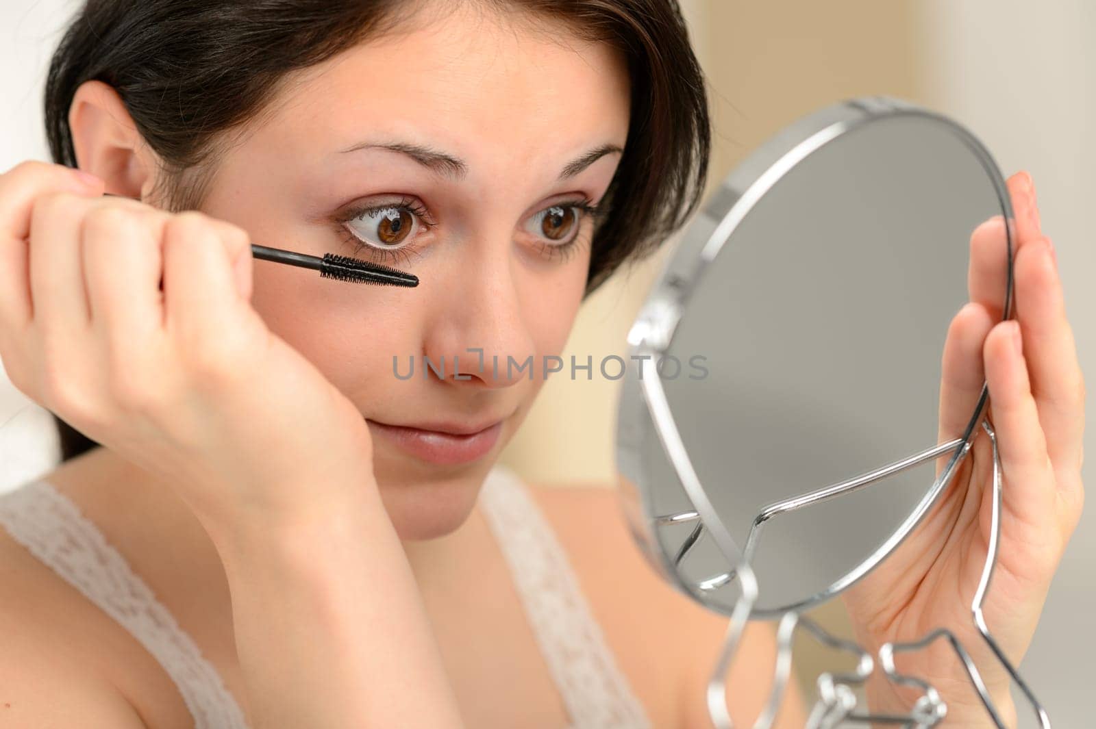 Attractive woman using mascara on her eyelashes holding handheld mirror
