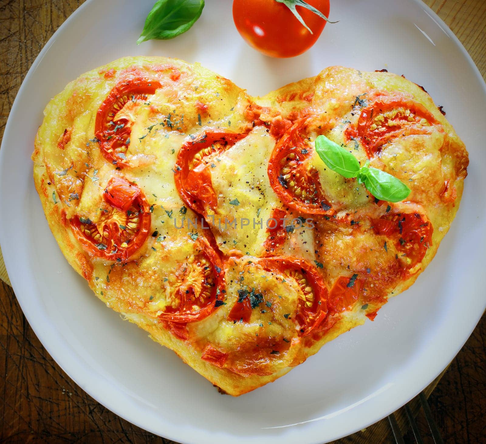 Overhead view of a romantic heart shaped Italian pizza topped with a vegetarian topping of golden melted cheese and tomato on a plain white plate. More pizza at my port.