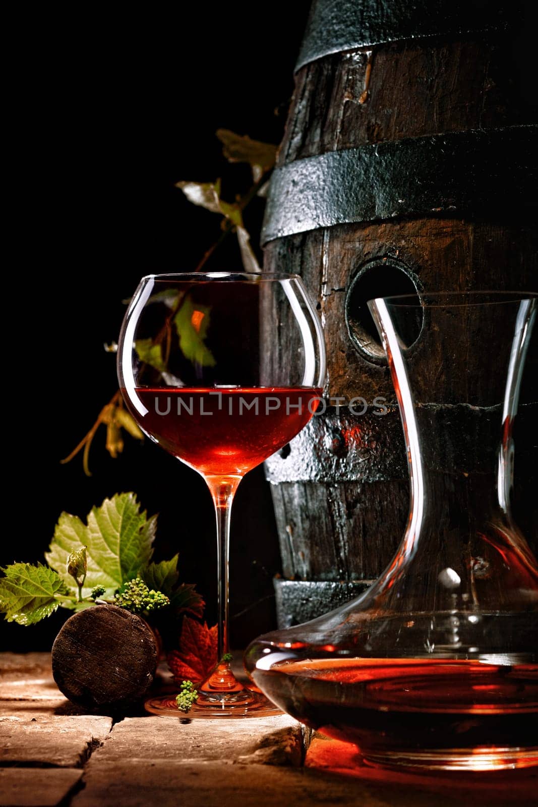 A glass of red wine next to a decanter with an oak cask in the background.