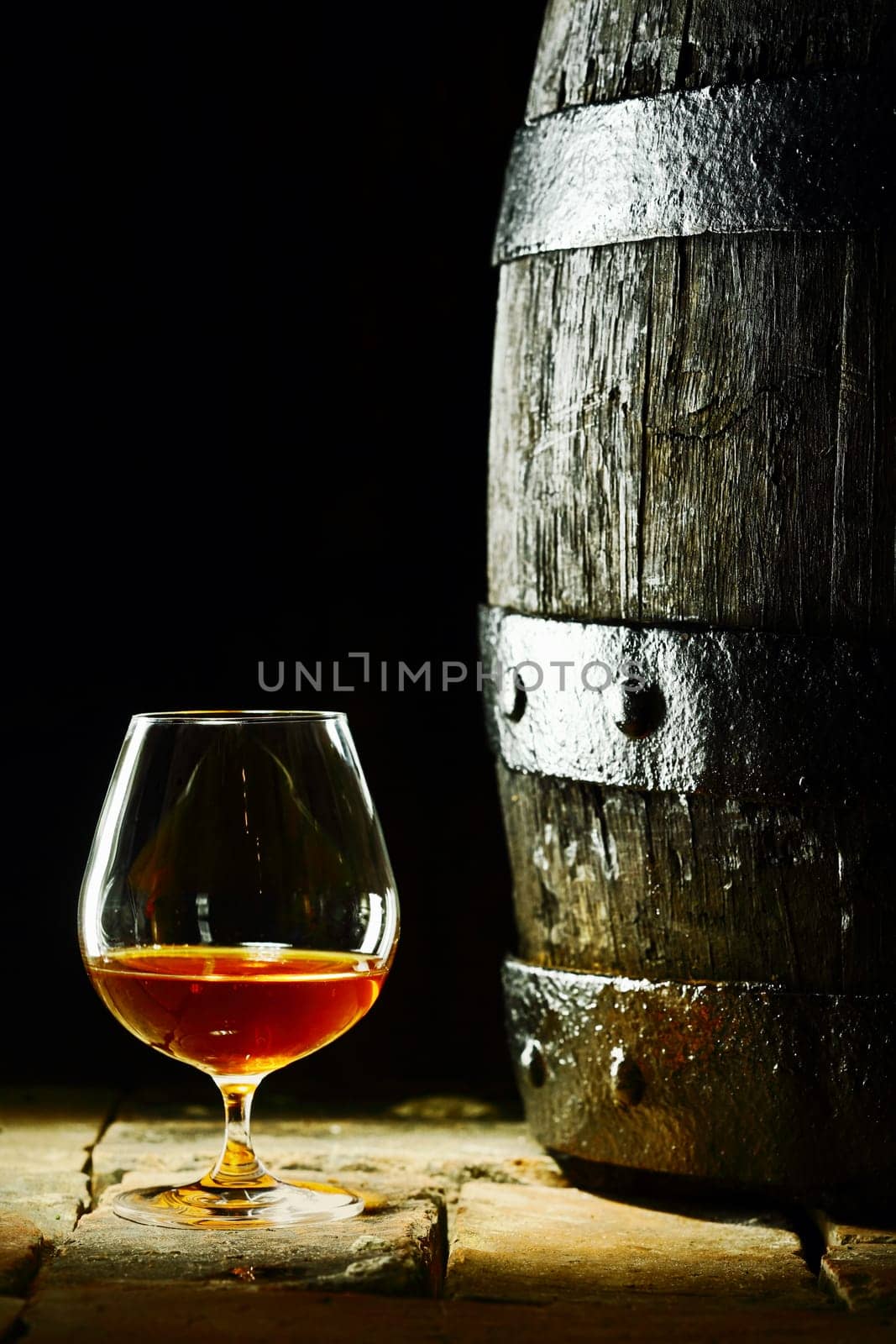 Cognac snifter with glowing golden cognac standing on old flagstones or tiles alongside an old oak barrel in a dark cellar, conceptual of aged matured cognac with copyspace