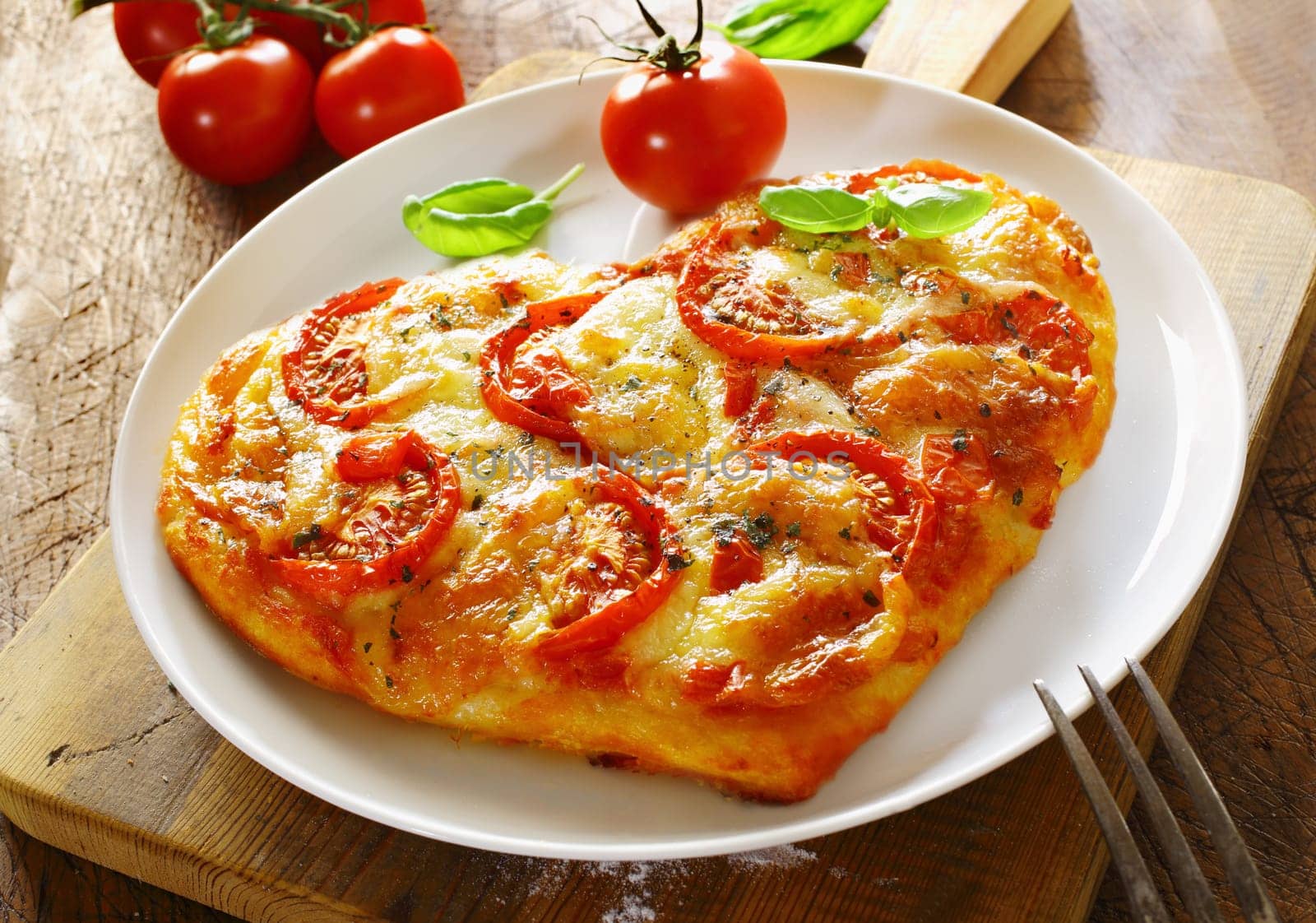 Delicious heart shaped Italian pizza with a topping of tomato and melted cheese served on a plain white plate with a fresh tomato and basil