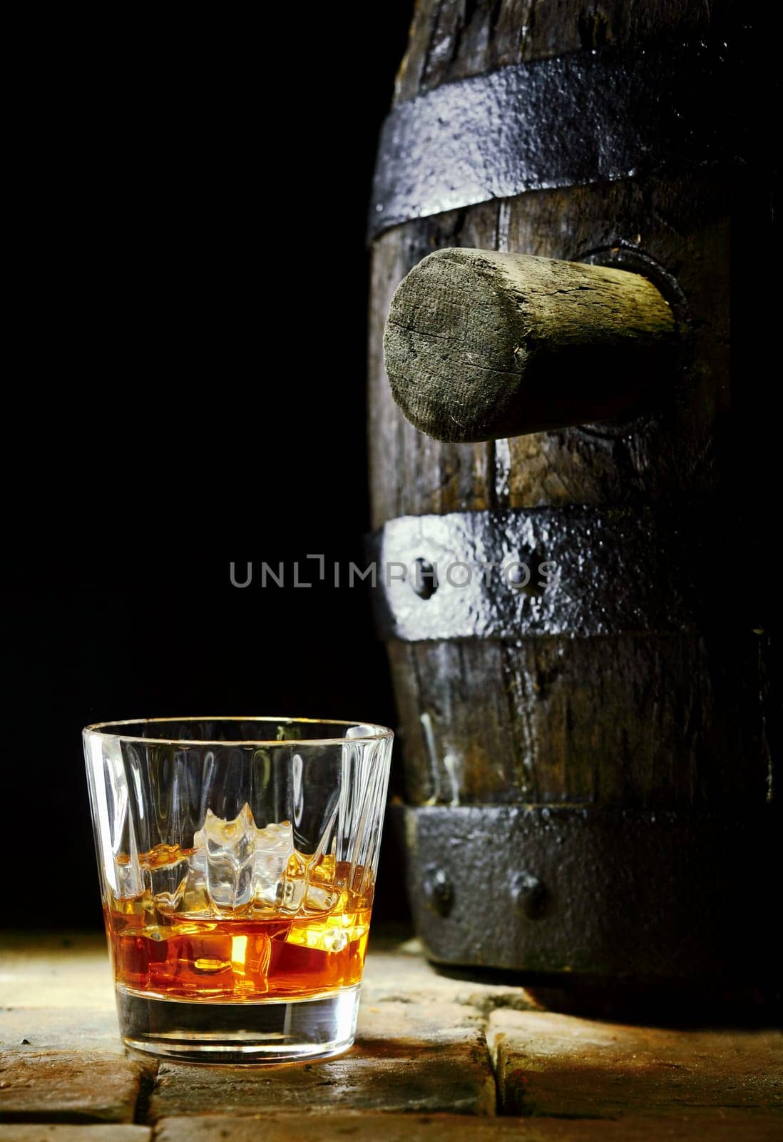 A glass of whiskey with ice and an oak barrel with it's stopper in.
