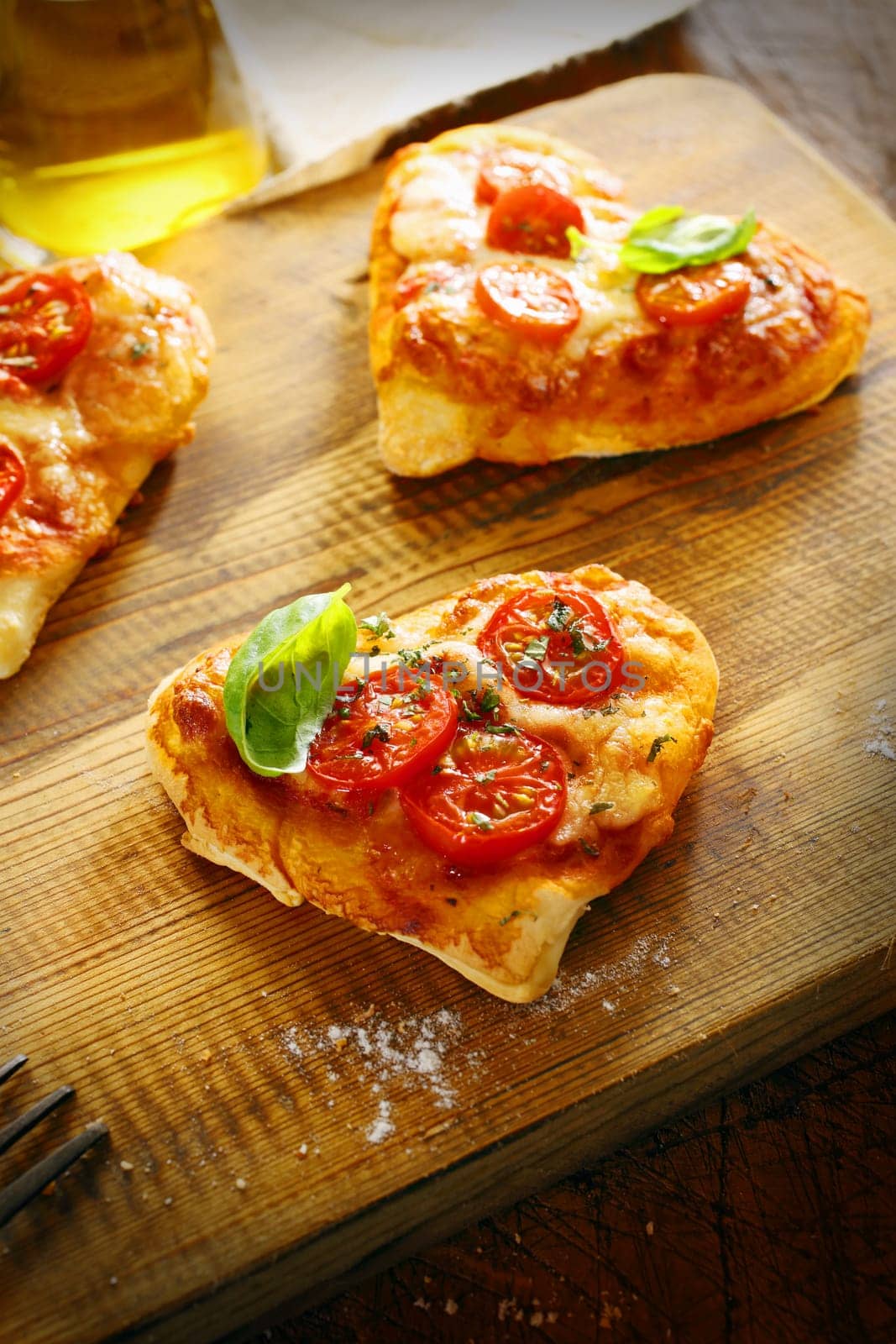 Small romantic heart shaped Italian pizzas with a freshly baked thick base topped with golden melted cheese and tomato and garnished with basil on a wooden board in the kitchen