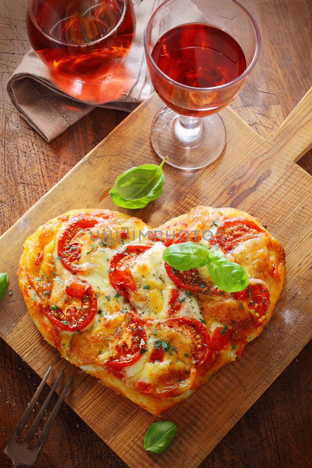 Overhead view of a tasty cheese and tomato vegetarian heart shaped pizza served on a wooden board with a glass of red wine