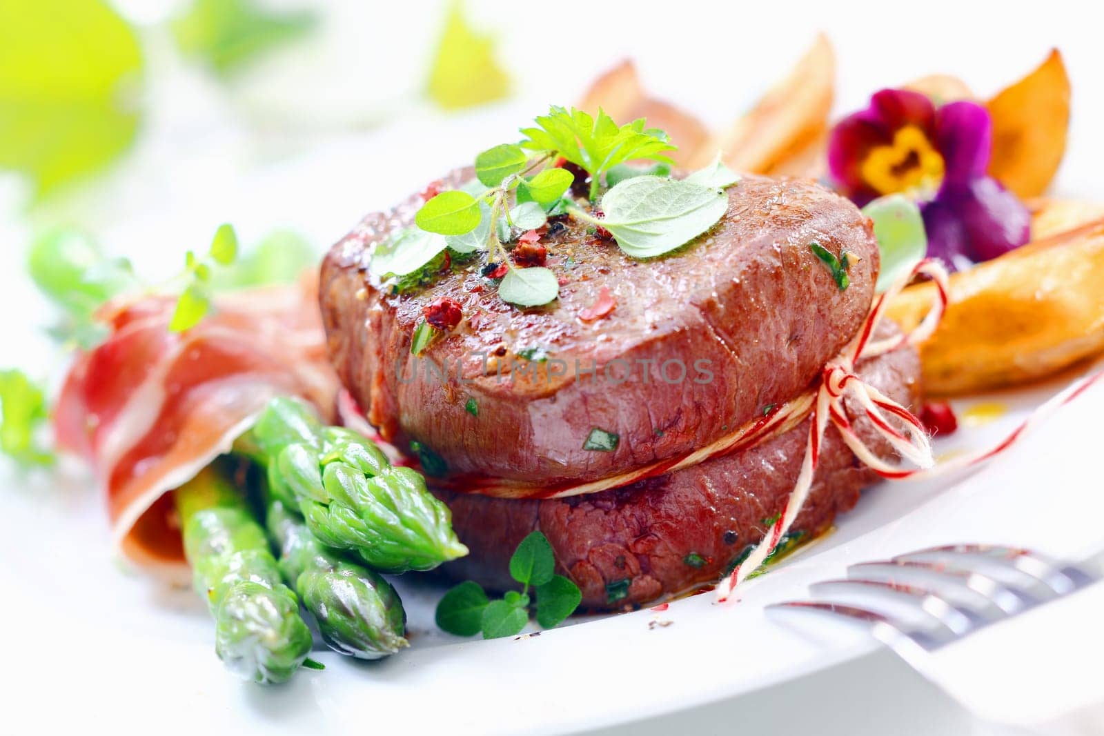 Thick juicy steak with fresh green asparagus spears wrapped in thinly sliced ham or bacon served garnished with herbs and crisp golden roast potatoes