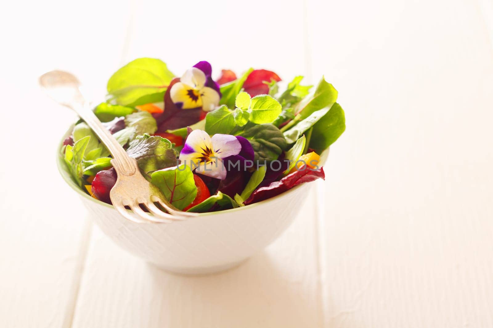 Healthy farm fresh herb salad with leafy greens and nasturtium flowers served in a white bowl with a fork on a wooden white table