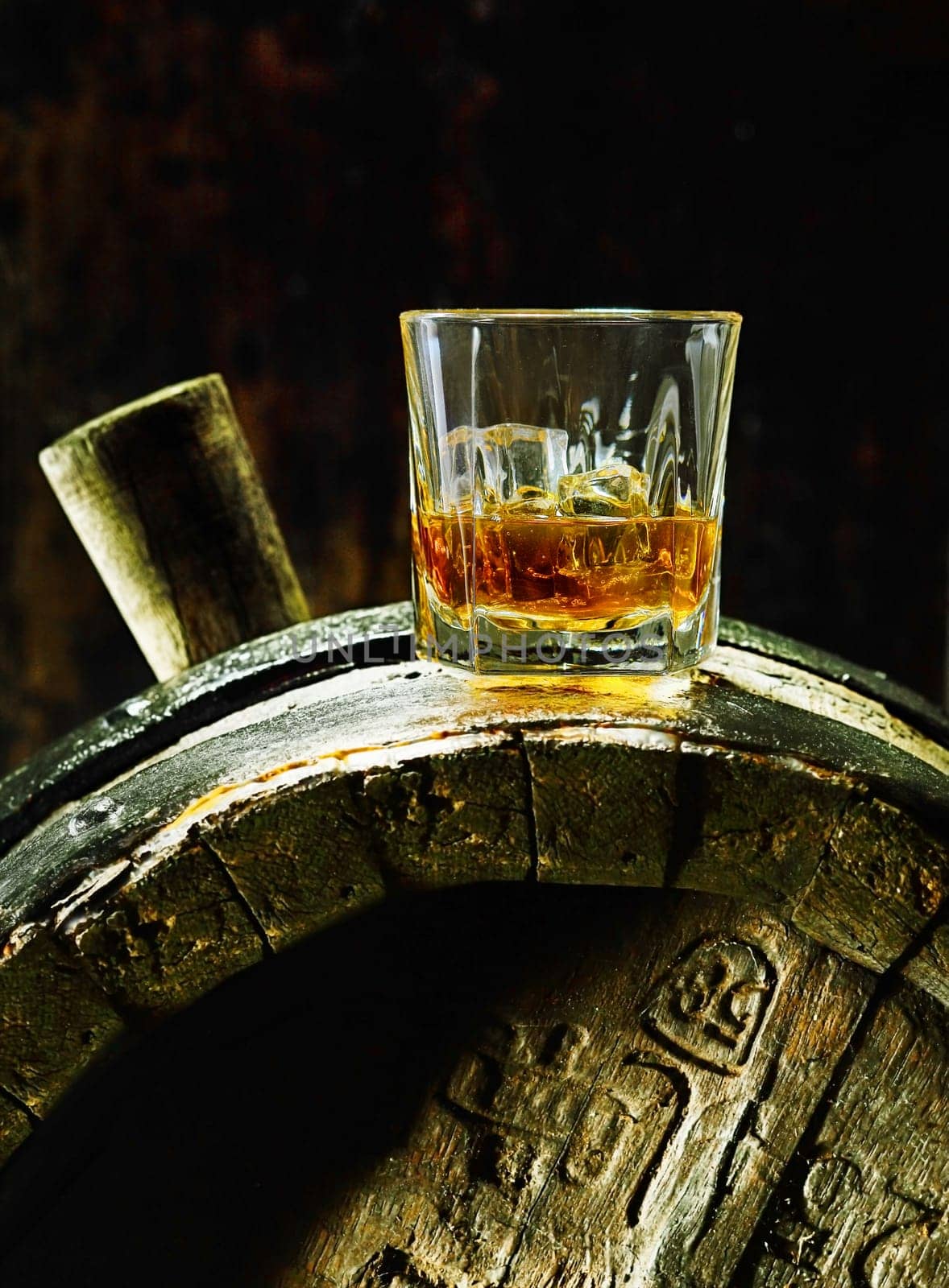 A glass of whiskey with ice on an old oak barrel using dramatic lighting.
