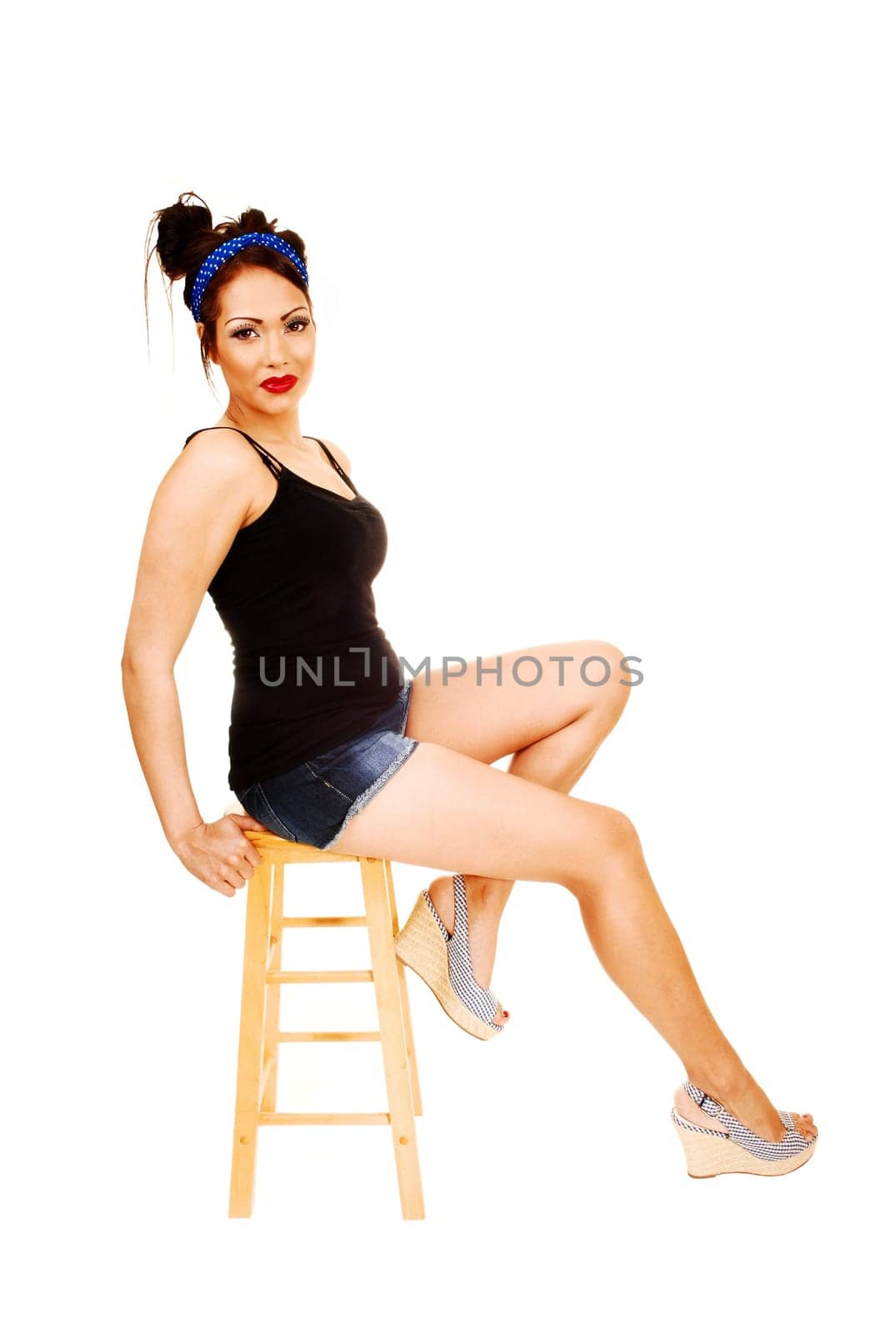 A young pretty woman in pin up style sitting on a chair with shorts and high heels and a blue headband in her hair, for white background.