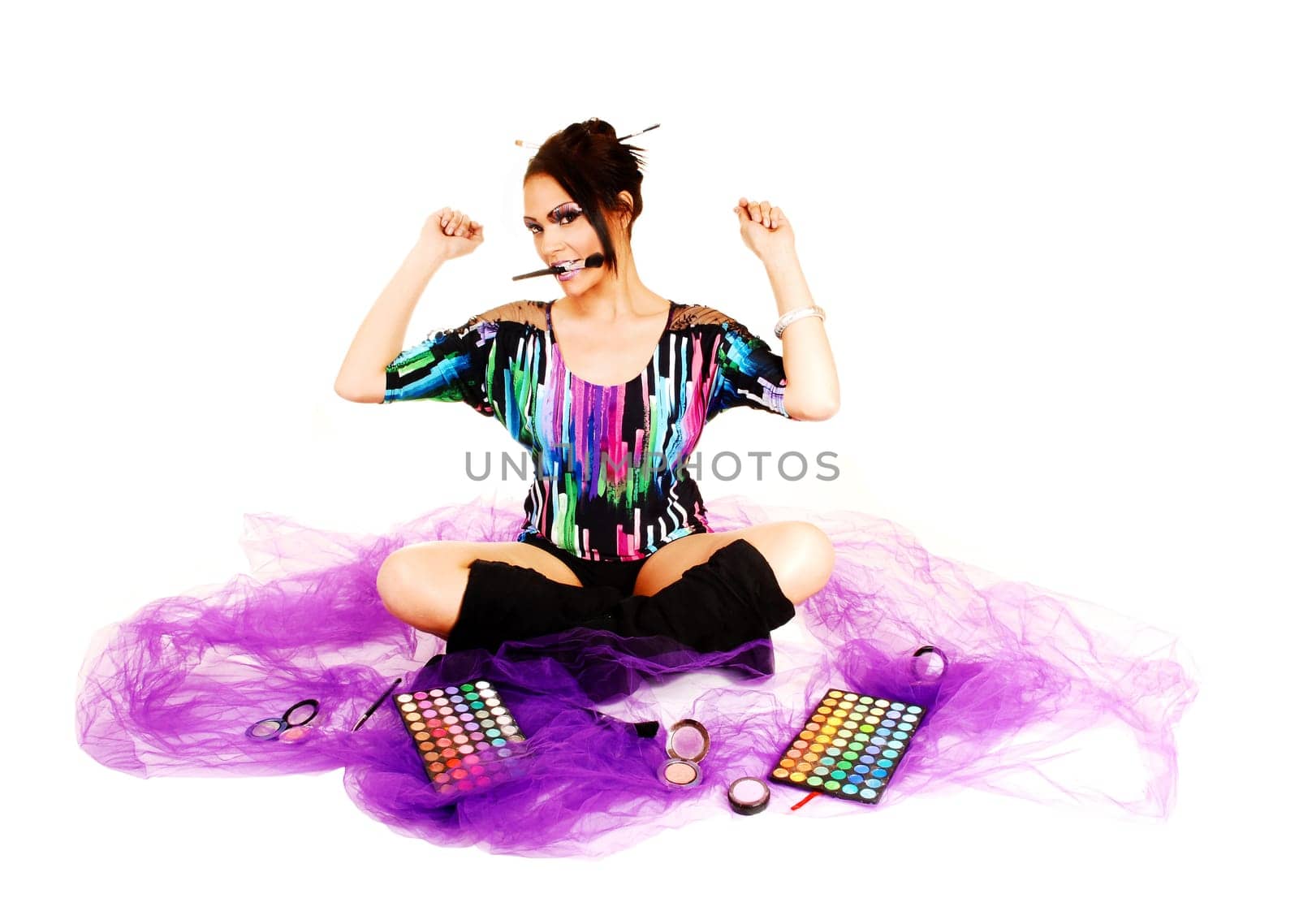 A young woman sitting on lilac twill on the floor with crossed legs and black long boots putting on her makeup, holding up her hands.