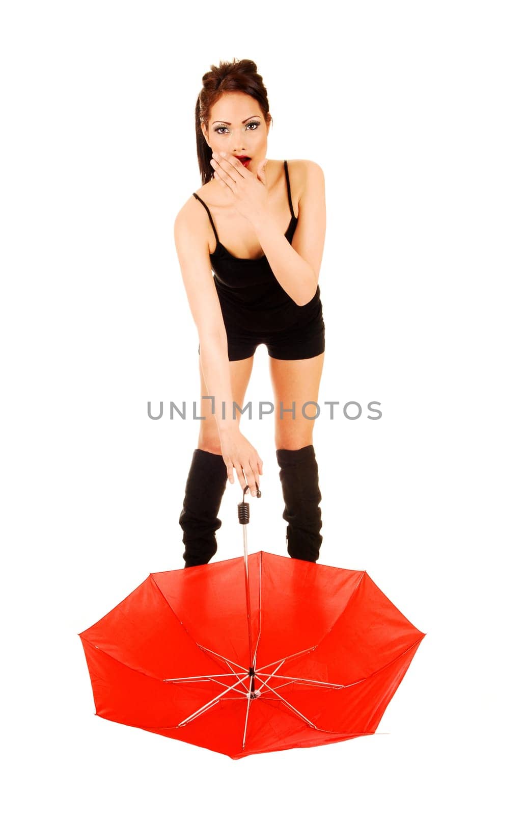 A young woman in black shorts and long boots standing with a red umbrella on the floor is looking very surprised for white background.