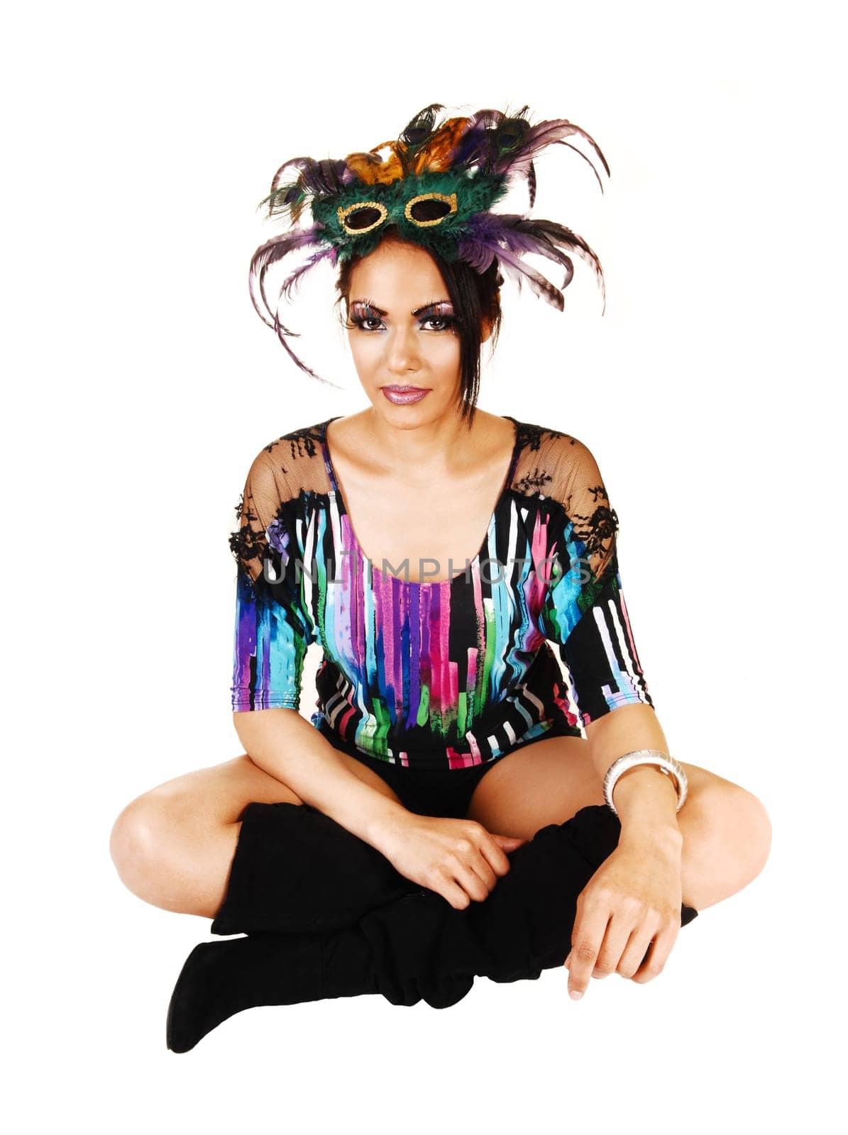 A young woman in a colorful blouse and black long boots sitting with closed legs on the floor with a father mask  on her forehead on white background.