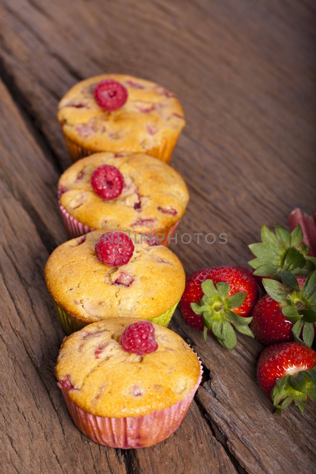Four strawberry,rhubarb,raspberry muffins in a row with fresh fruits in the background