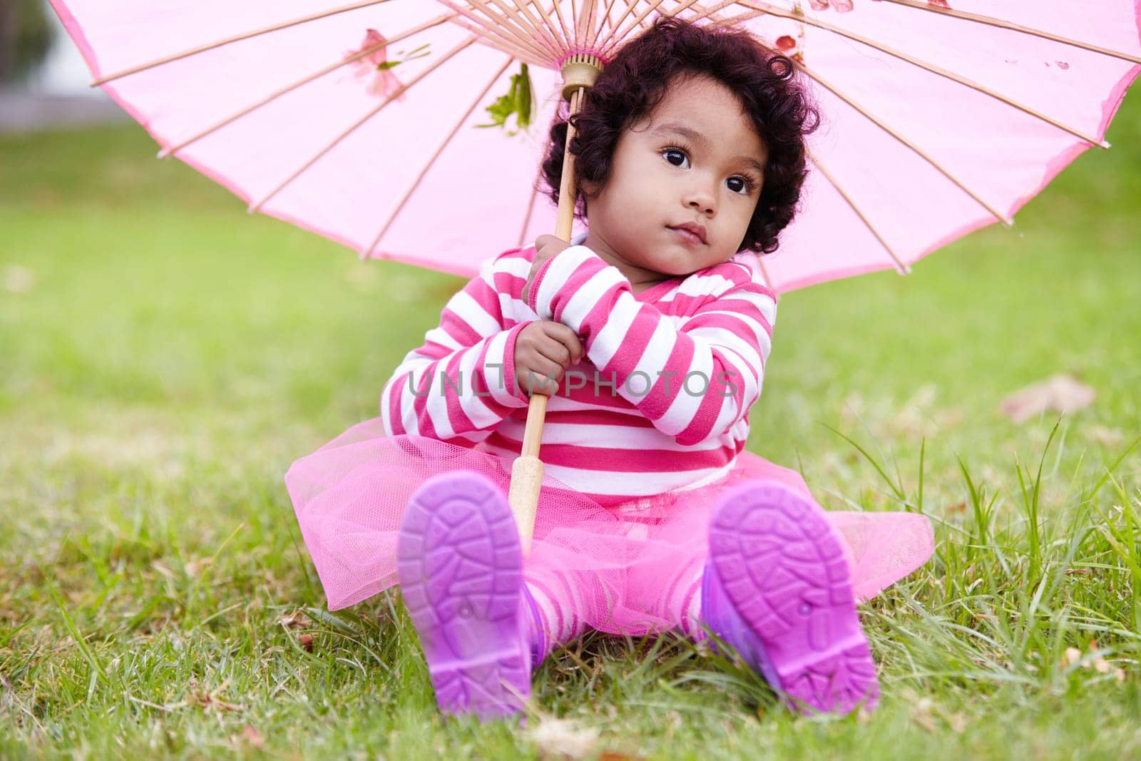 Umbrella, cute and girl child in a garden sitting on the grass on summer weekend. Adorable, playful and young kid, baby or toddler with curly hair playing on the lawn in outdoor field or park