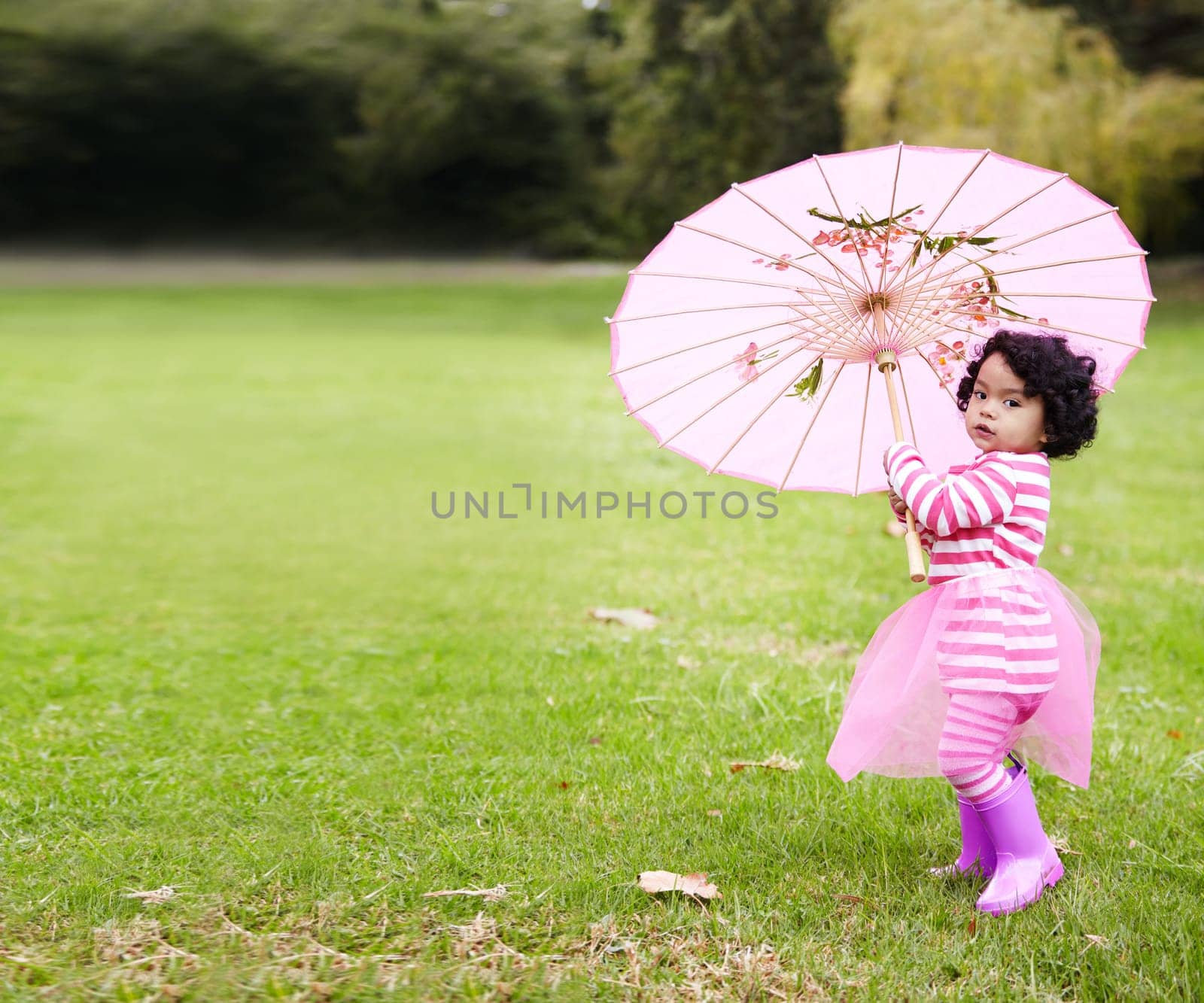 Umbrella, walking and girl child in a garden walking on the grass on summer weekend. Adorable, playful and young kid, baby or toddler with curly hair playing on the lawn in outdoor field or park