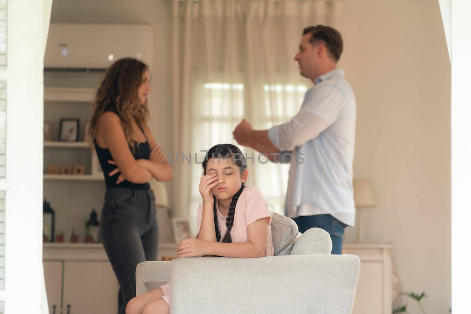 Annoyed and unhappy young girl sitting on sofa trapped in middle of tension by her parent argument in living room. Unhealthy domestic lifestyle and traumatic childhood develop to depression.Synchronos