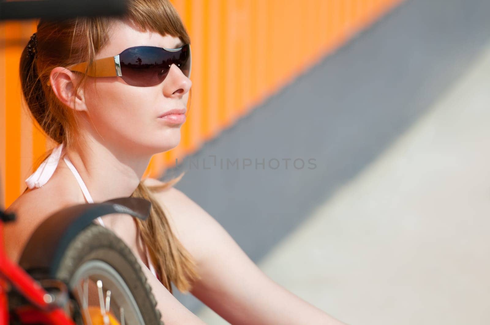 Beautiful young woman sitting near the wall, wearing sunglasses. Fragments of her bicycle in front of her.