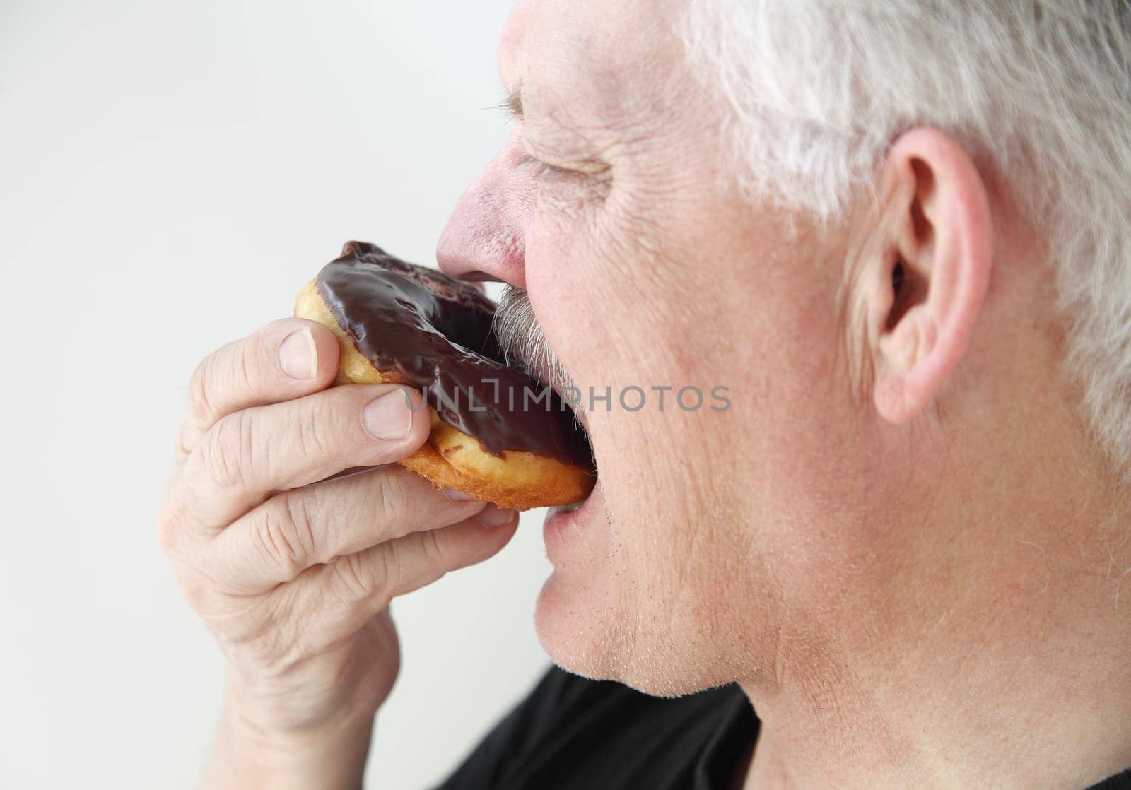 profile view of senior man biting into a chocolate glazed doughnut