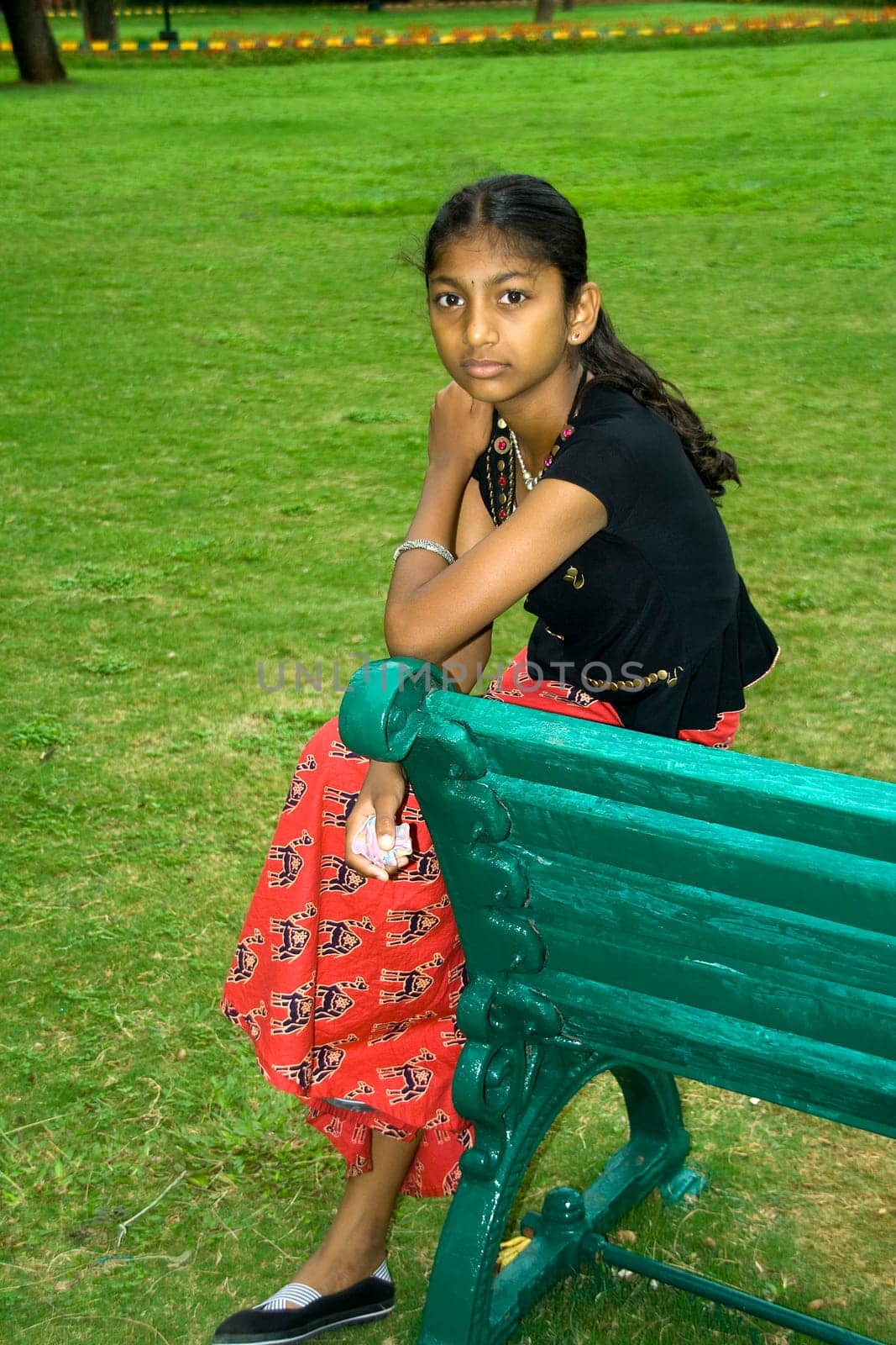 Girl sitting on the edge of wooden bench placed on green turf in a garden