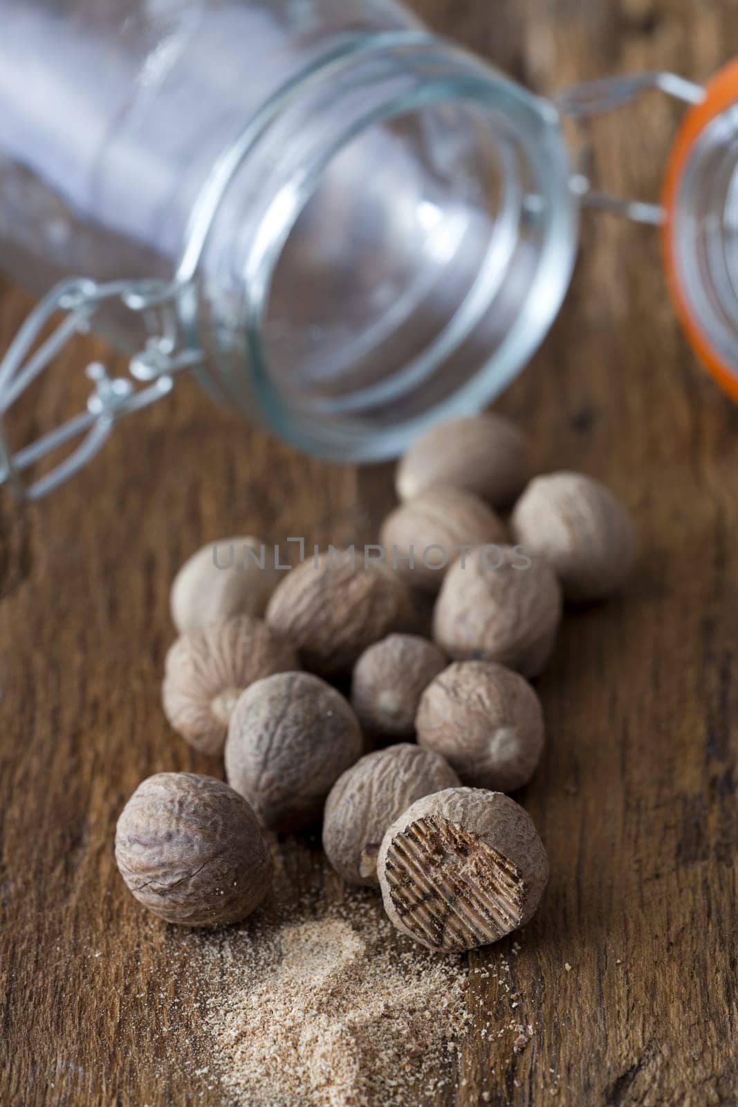 Numerous nutmegs lie in front of an empty spice jar on a rustic wooden surface