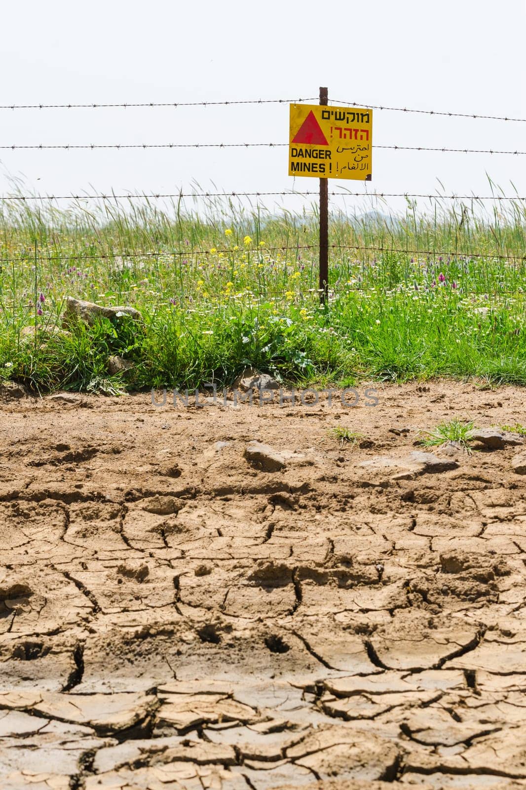 Cracked dried ground against green minefield with razor wire and warning sign