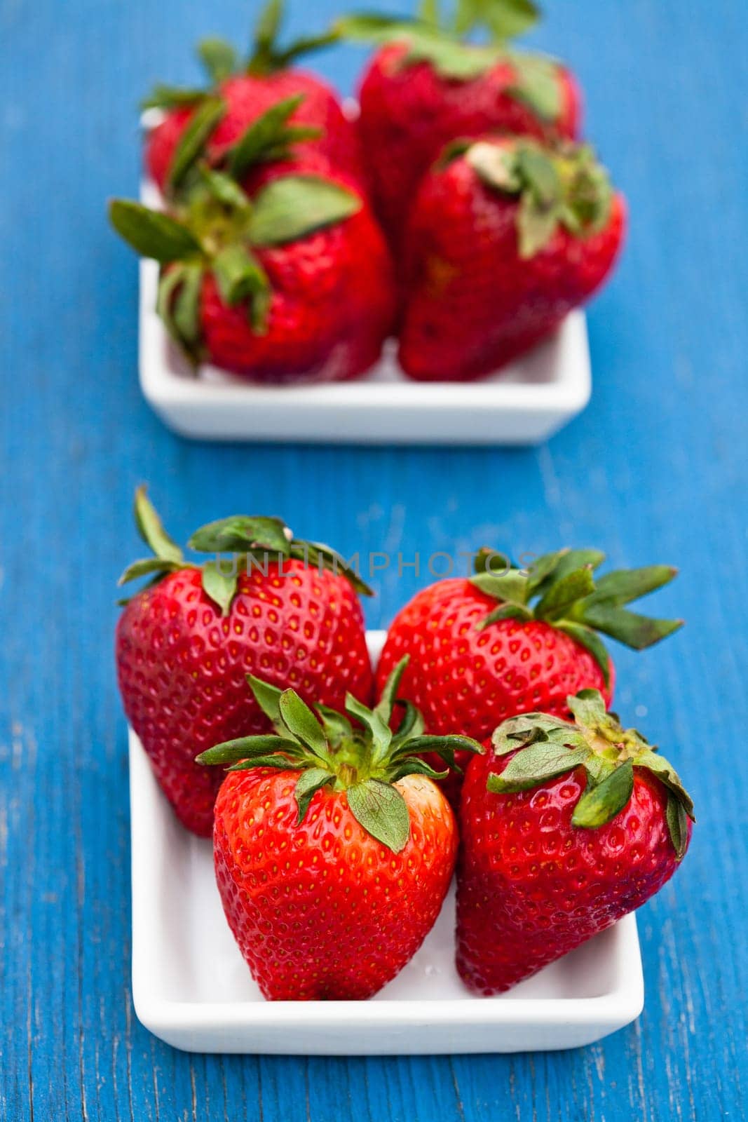 Closeup of large fresh strawberries in two white dishes on light blue wooden background