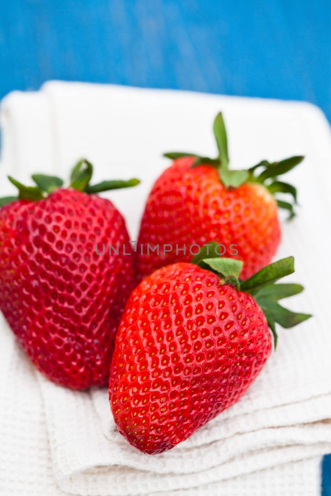 Closeup of three large fresh strawberries resting on white cloth against light blue wooden background