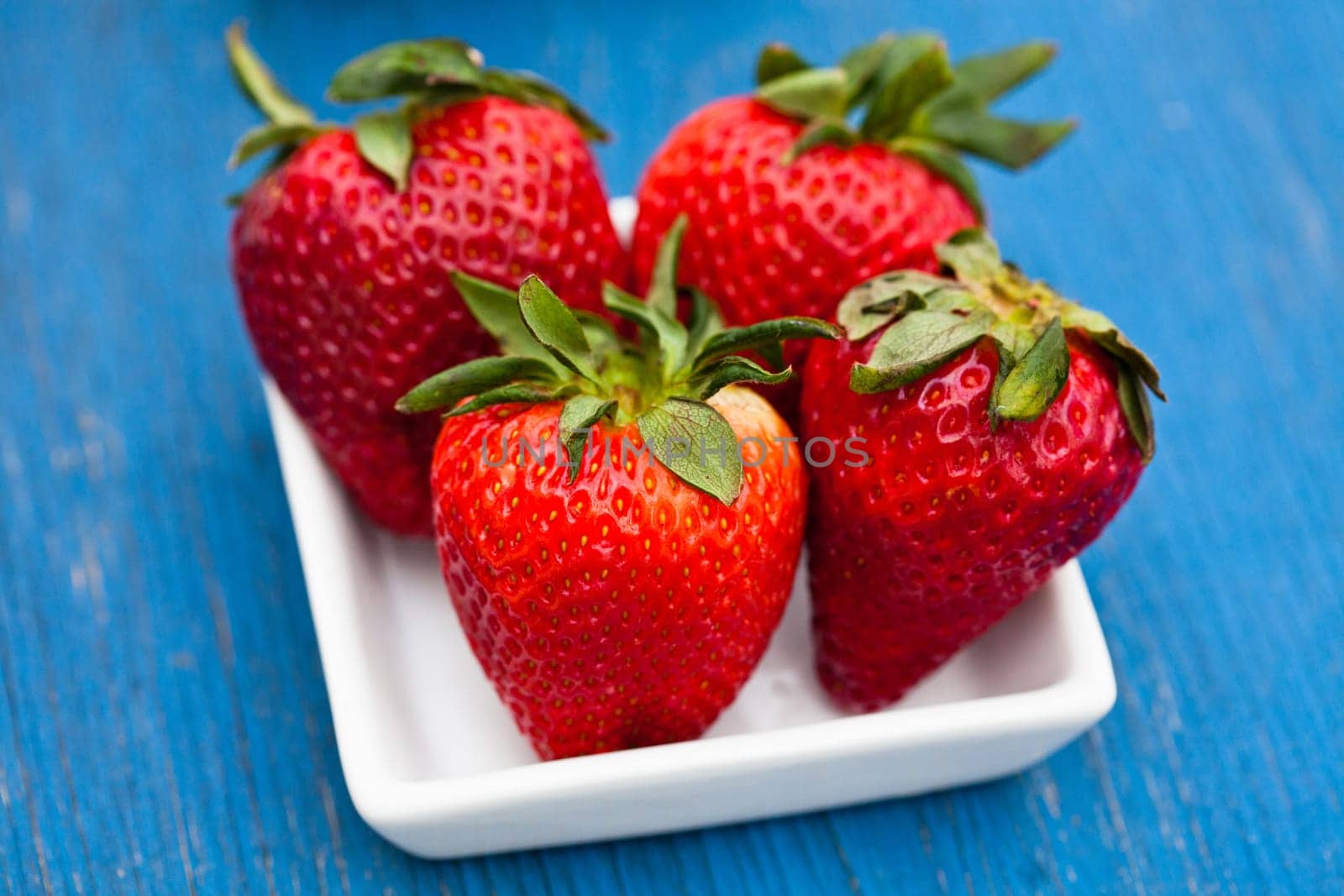 Closeup of four large fresh strawberries in white dish on light blue wooden background