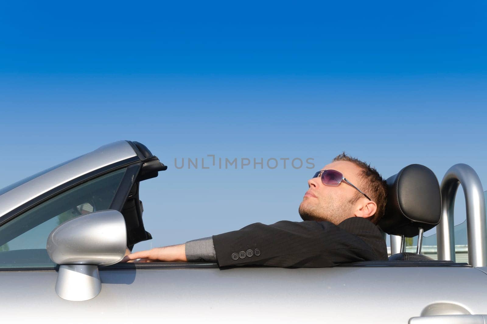 Young man in a dark suit and sunglasses sitting relaxed in his silver convertible sports car against a blue sky.