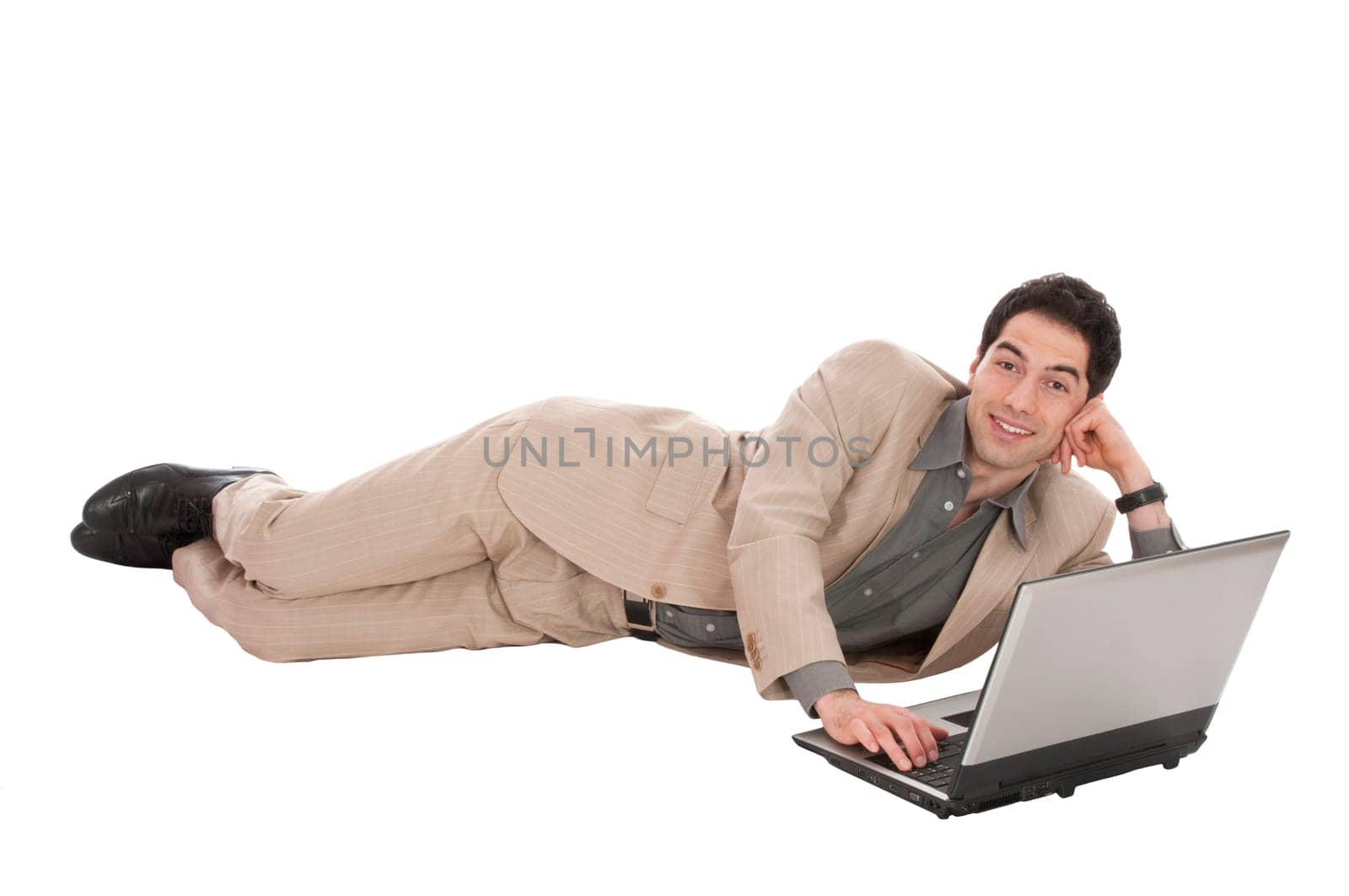 Young businessman in light suit lying on the floor and working on laptop, isolated against white background, laughing.