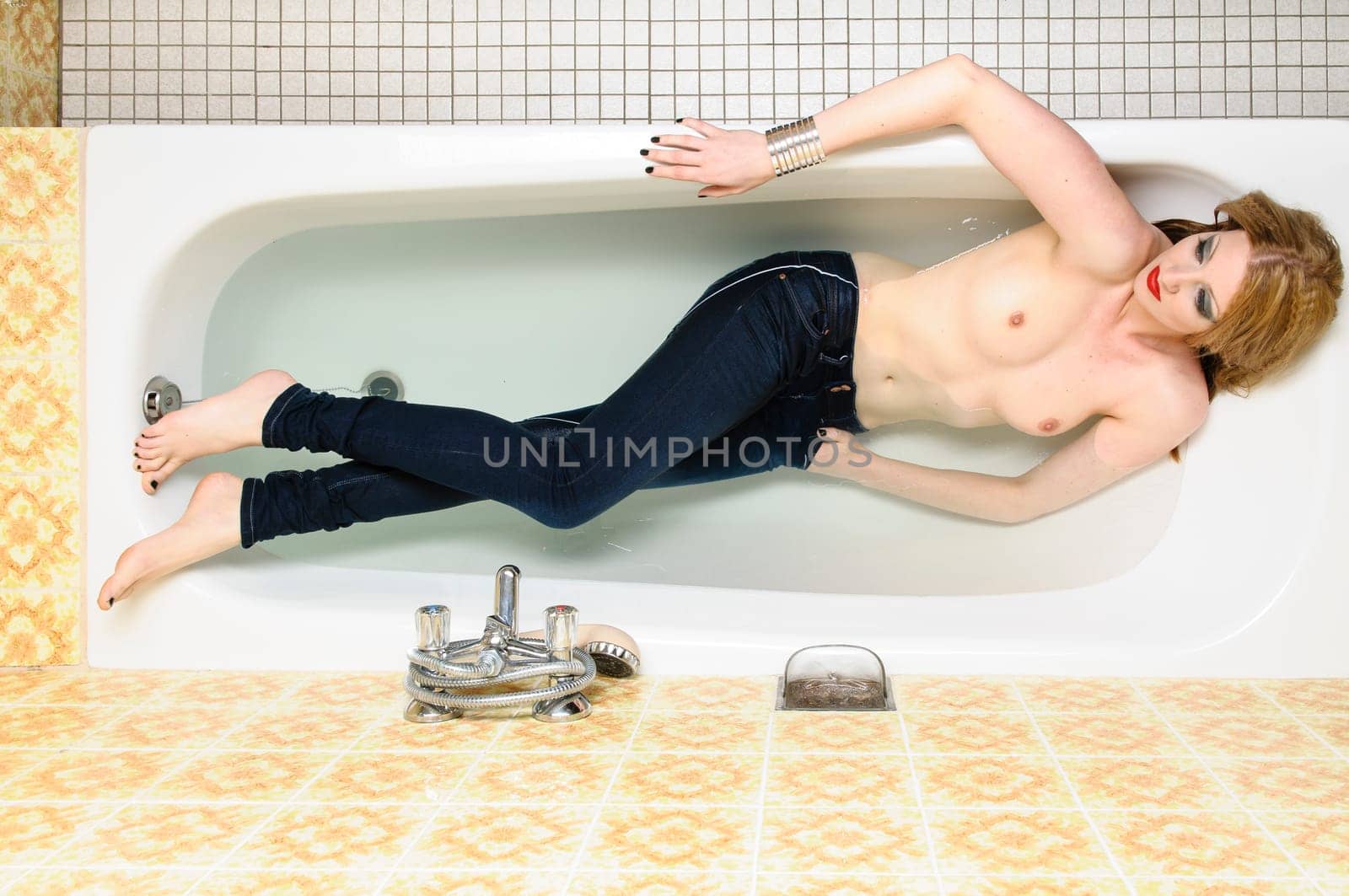 Beauty blond young woman lying with her bluejeans in the bathtub in water.