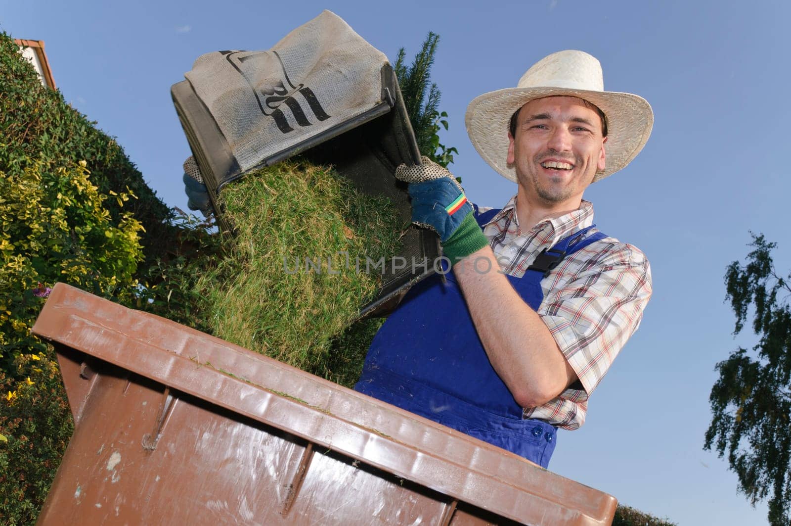 Man with blue overalls and a plaid shirt and straw hat standing in the garden next to the lawn mower and the compost bin filled with grass clippings.