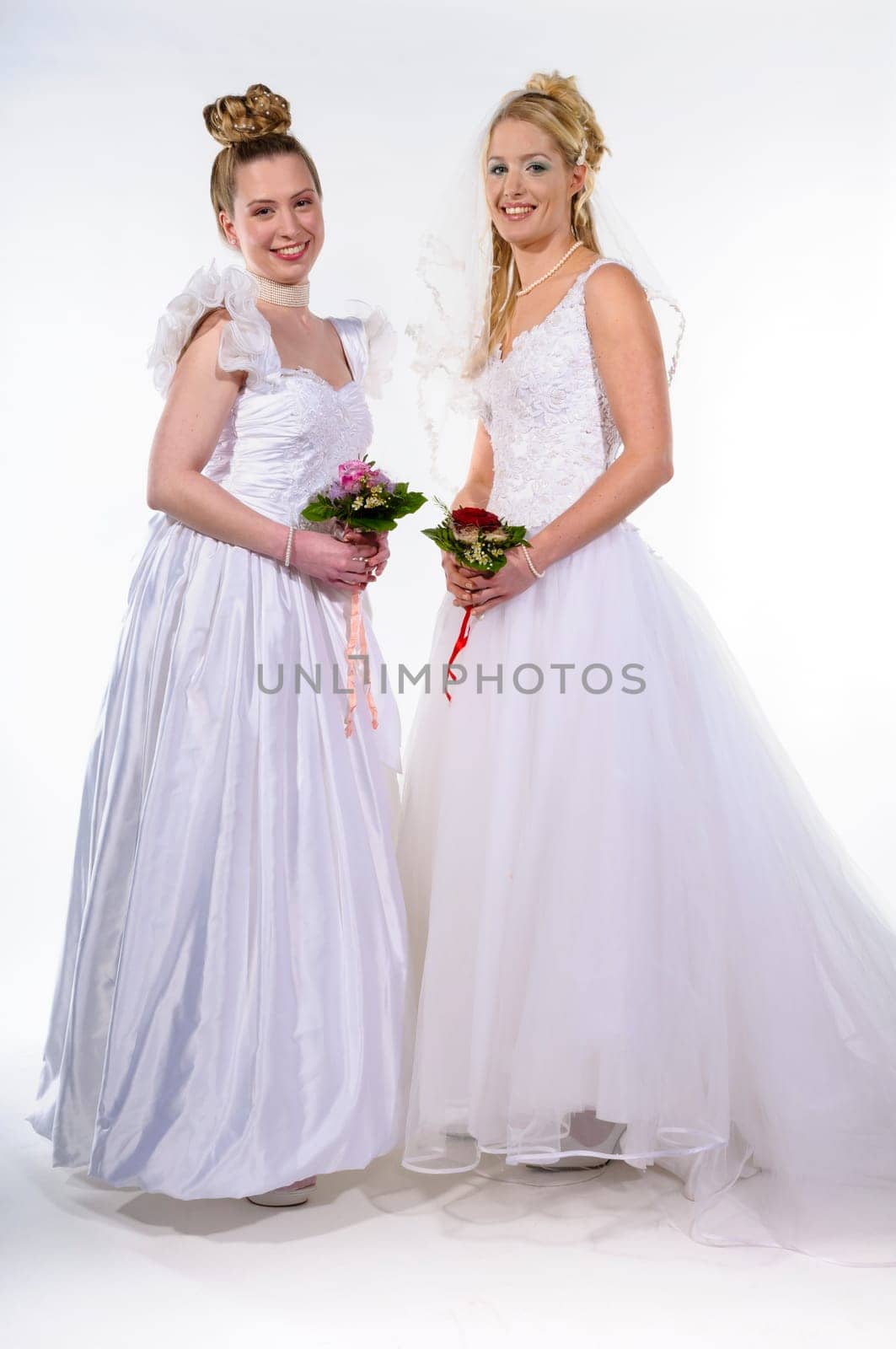 Two young women wearing bride dresses und holding their bride bouquets, against white background. Smiling in to camera