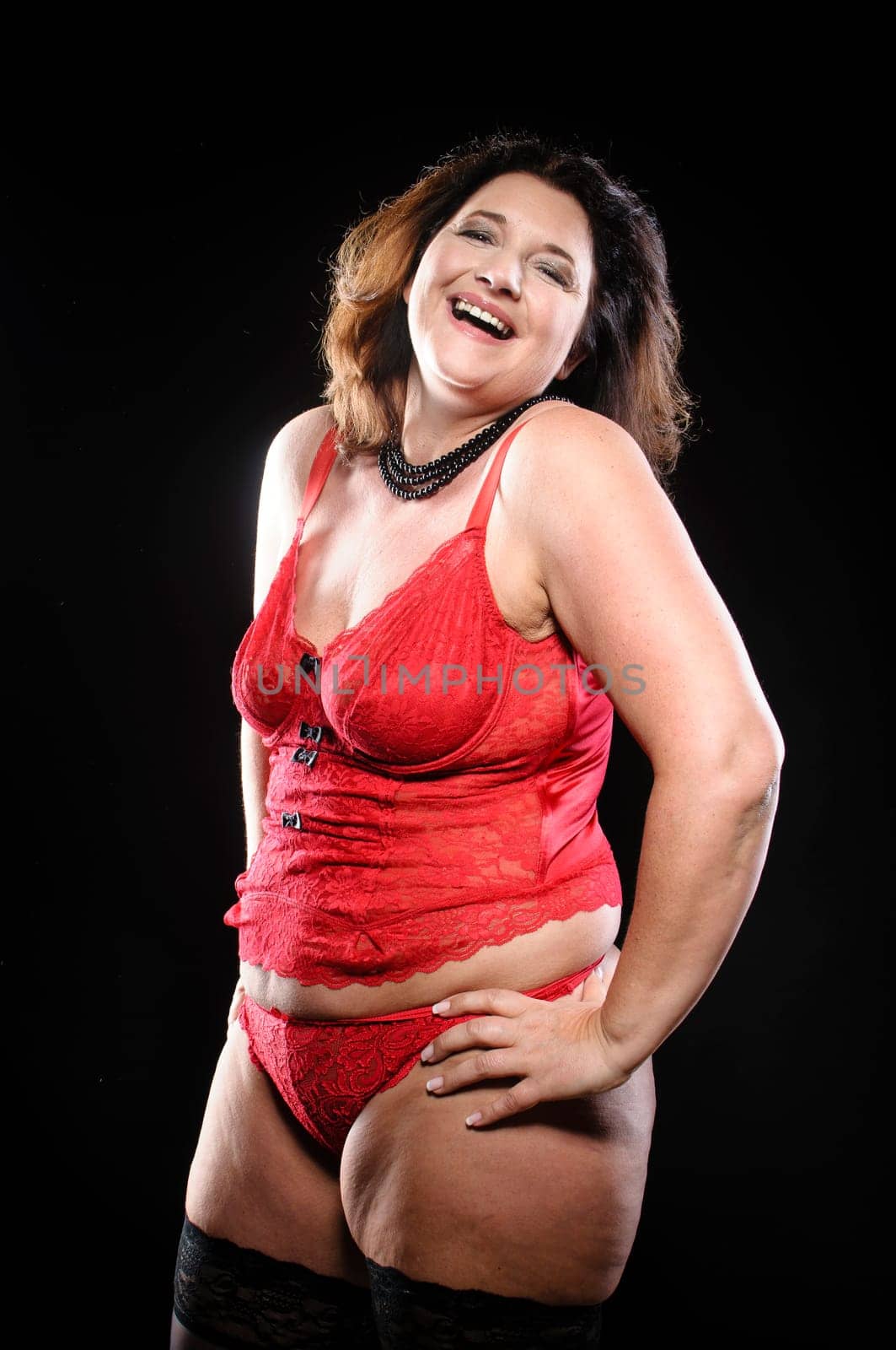 Attractive adult plus size model wearing red lingerie against black background and laughs.