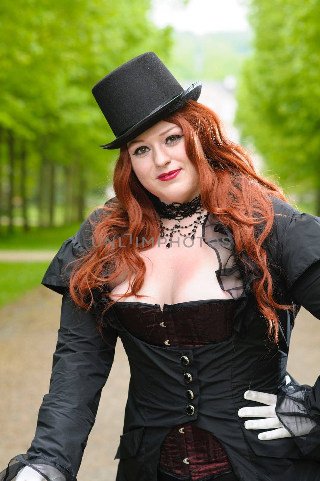 Young plus size model wearing fance dress with red corset, black overcoat and top-hat, standing in a park.