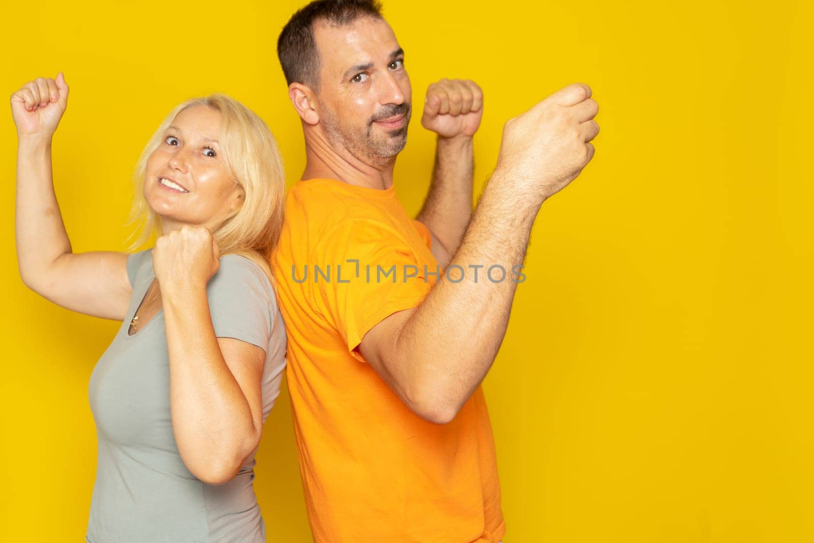 Back to back happy caucasian couple in their 40s celebrating victory with hands in the air on yellow studio background