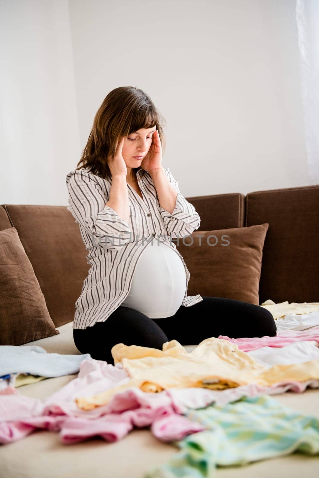 Pregnant woman with headache above baby clothes at home on sofa