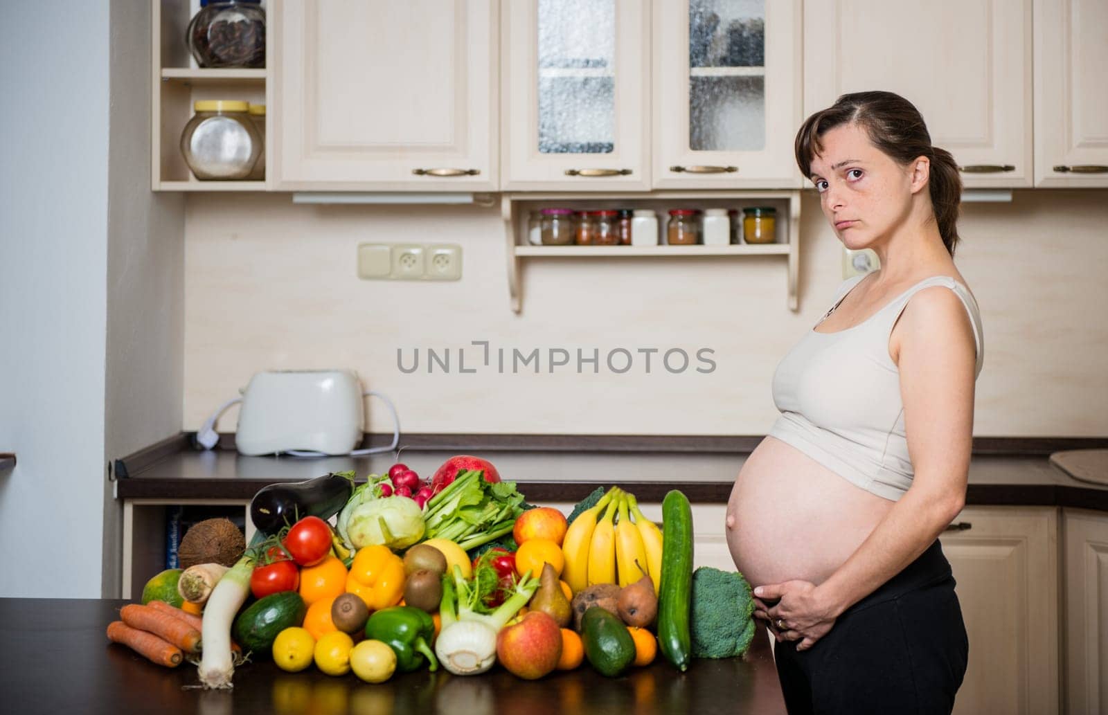 Unhappy pregnant woman with abundance of vegetables and fruits in kitchen