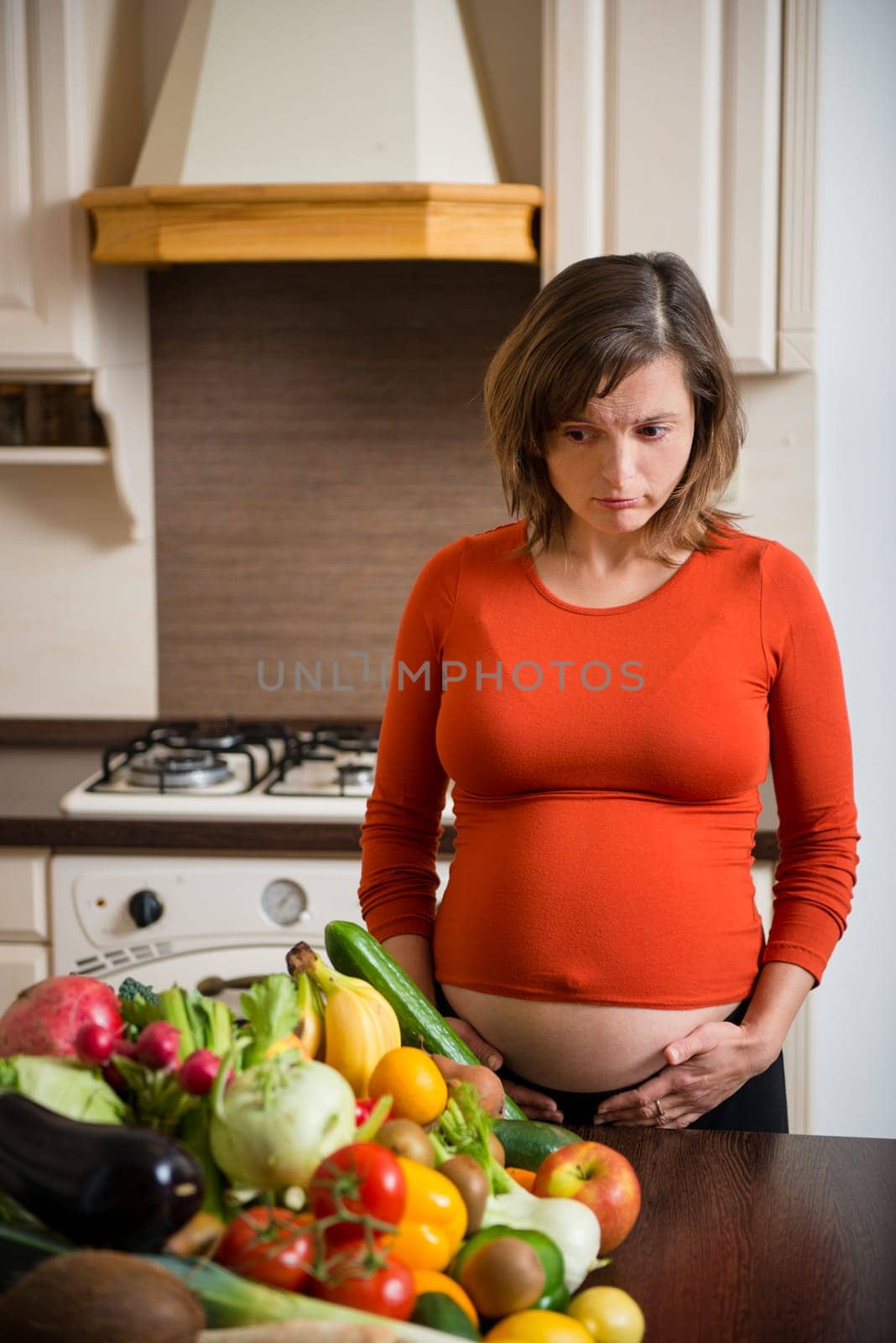 Unhappy pregnant woman with abundance of vegetables and fruits in kitchen