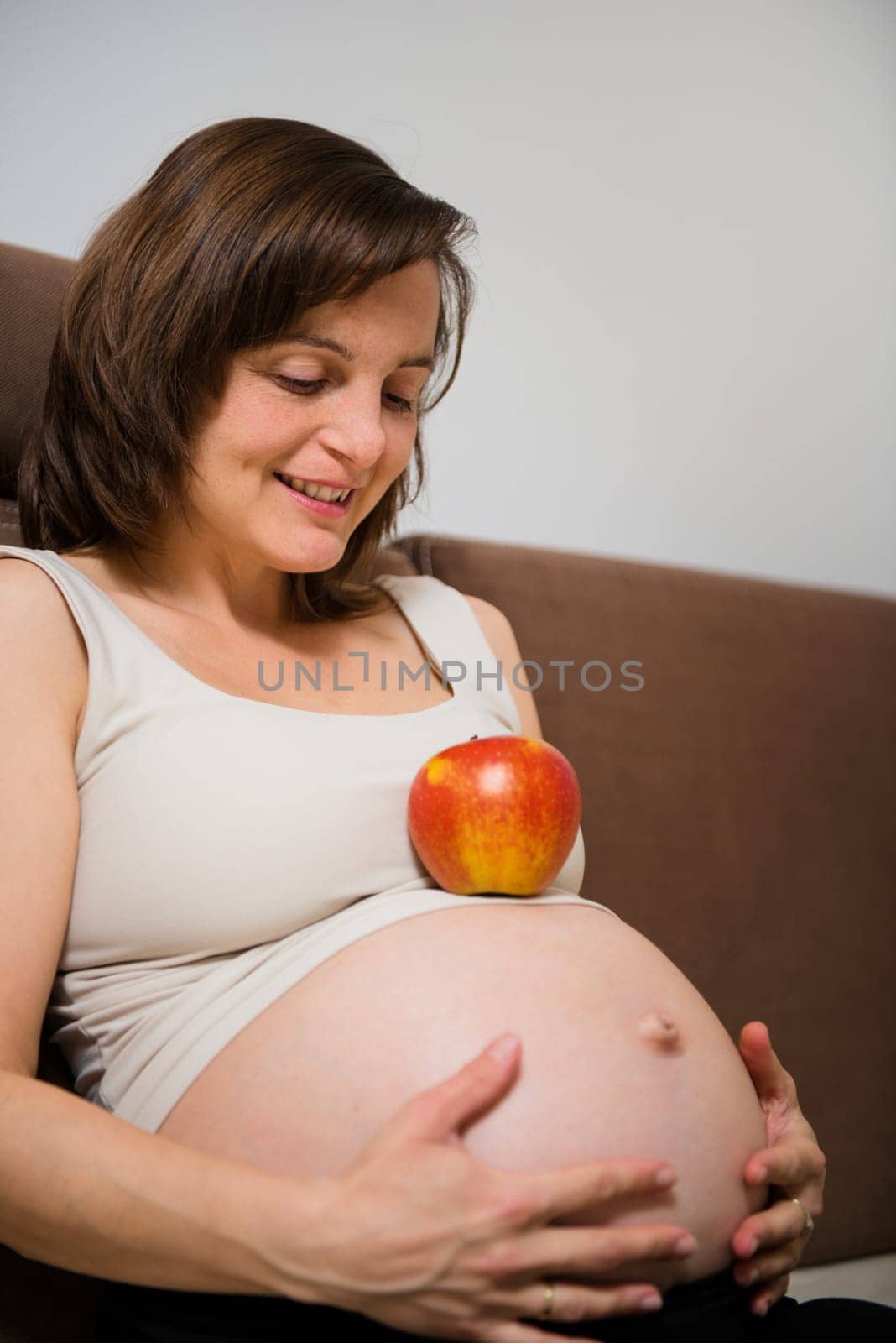Lifestyle portrait of smiling pregnant woman with apple on sofa