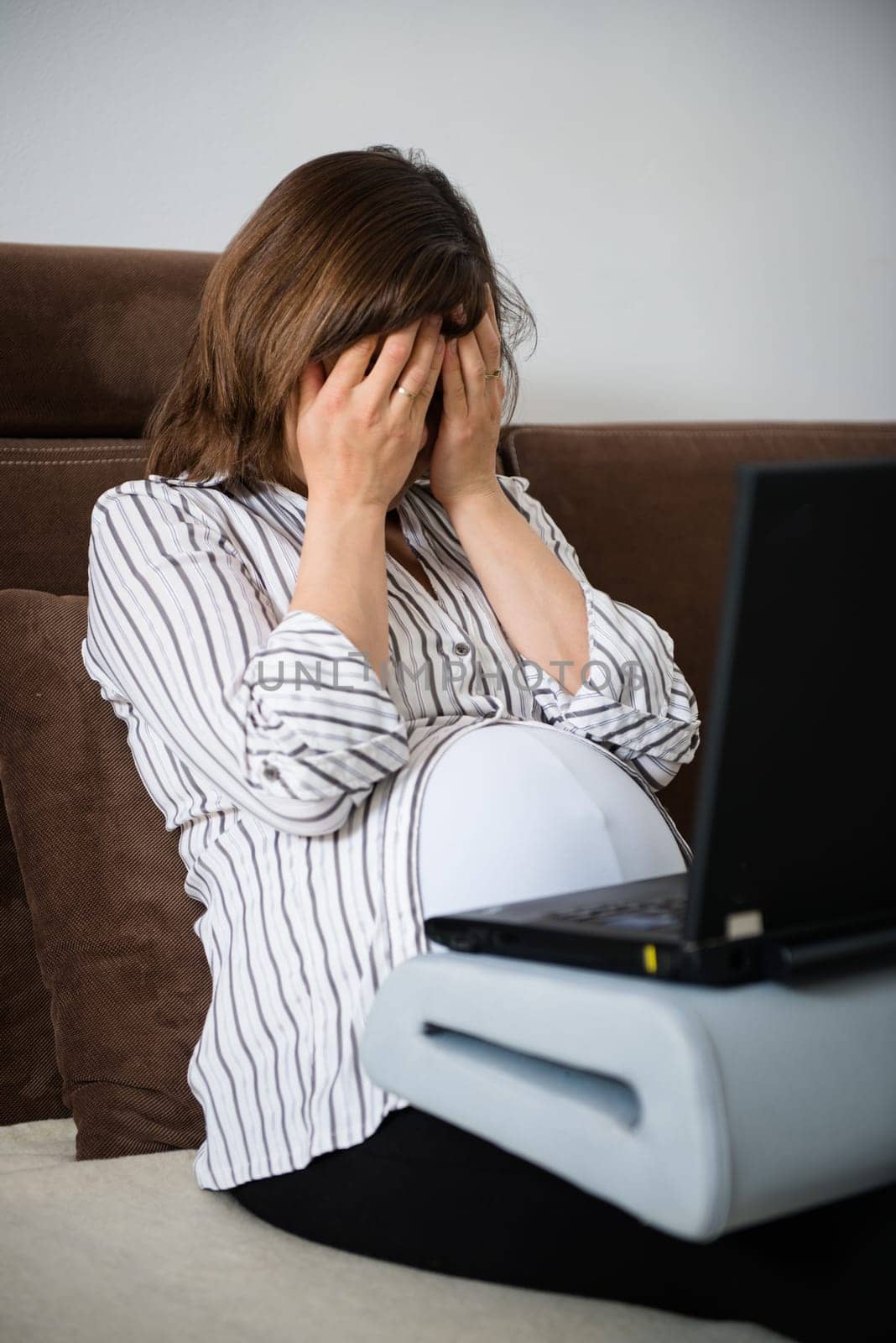 Pregnant woman with headache working on laptop at home on sofa
