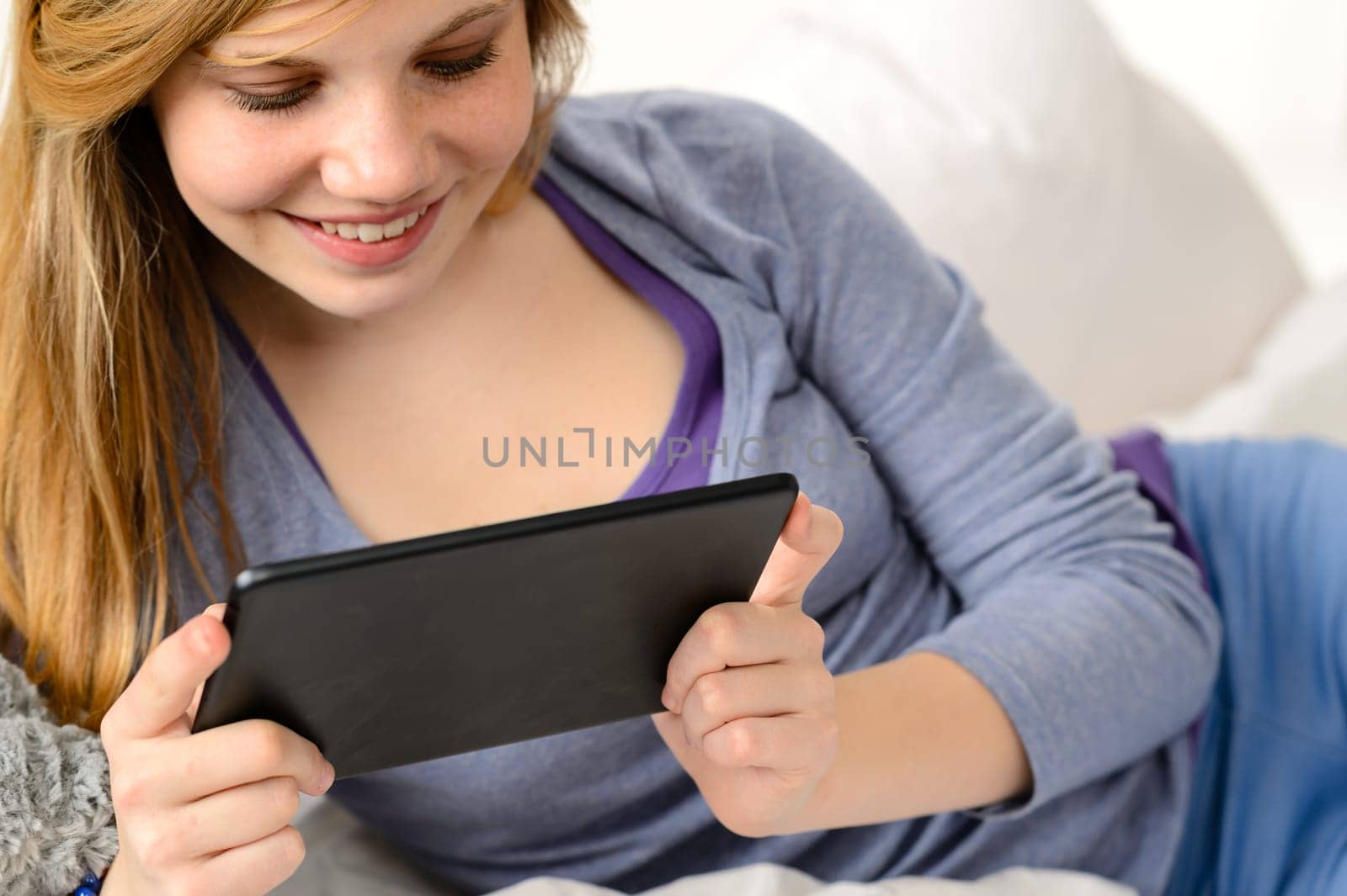 Friendly teenage girl reading on digital tablet lying in bed