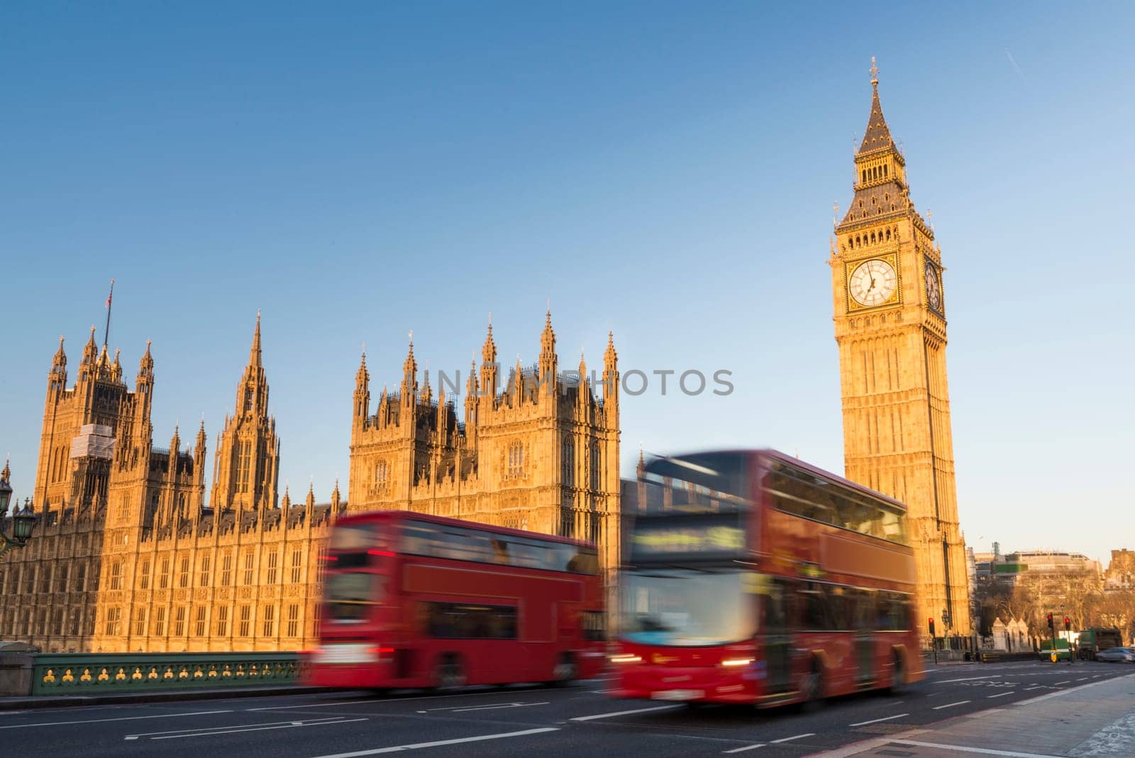 North face of Big Ben with houses of parliament, in London, UK, and motion blurred red double decker buses passing by.