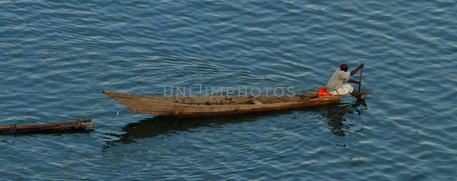 A lone African man paddles his long boat across a lake while pulling some logs.