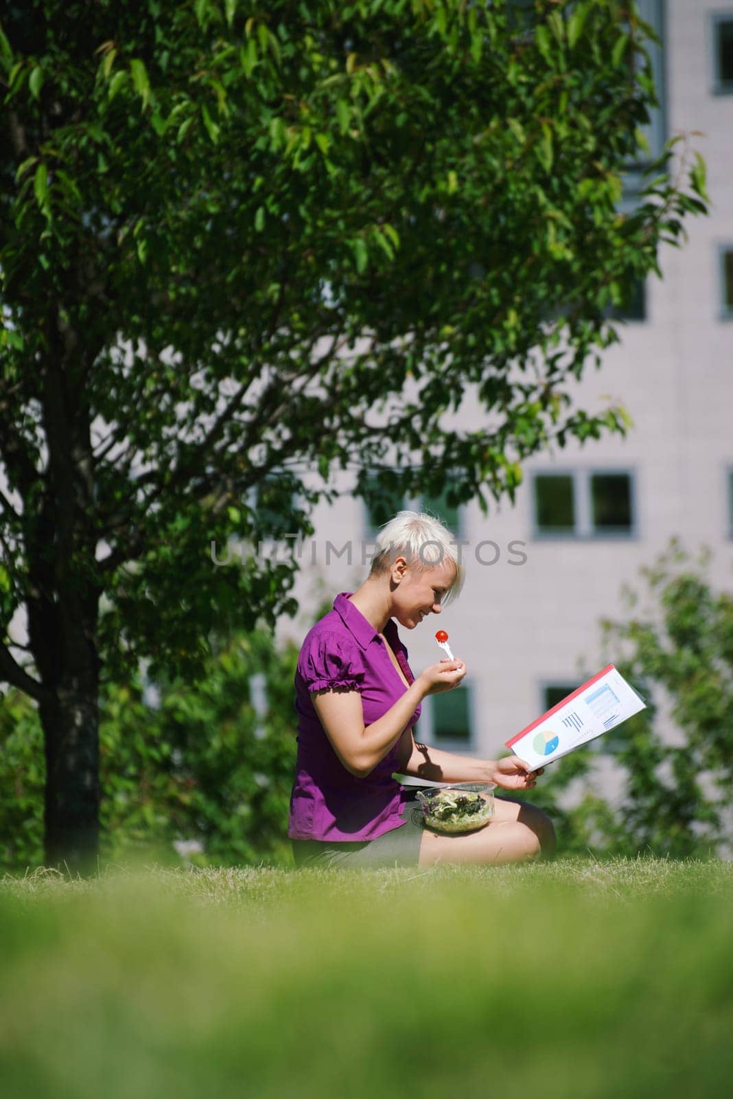 young business woman sitting on grass in park, eating salad for lunch and reading paperwork