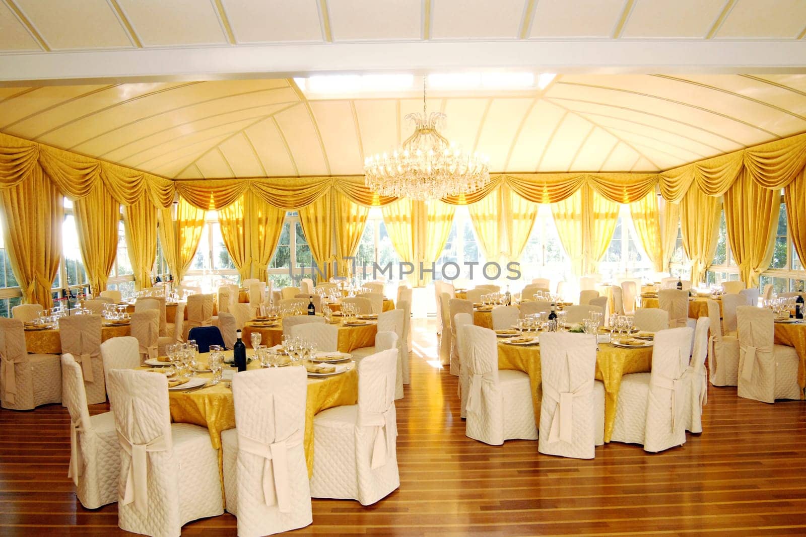 Luxury wedding or function venue interior with tables and table settings highlighted in a rich gold and multiple windows giving a bright airy light